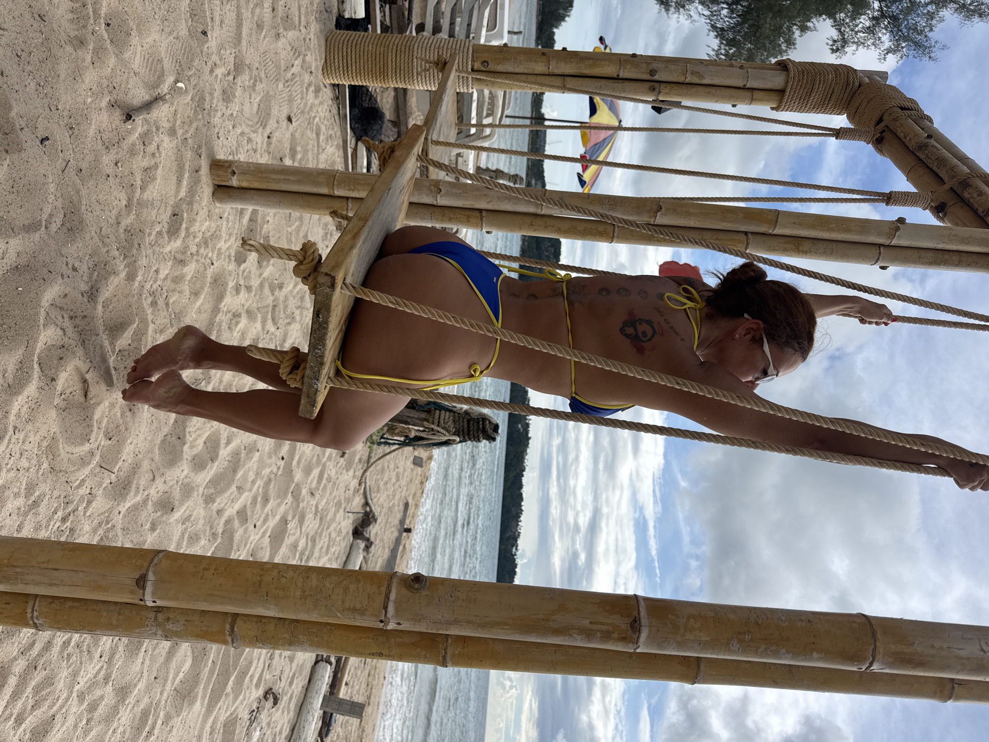 A woman wearing a blue and yellow bikini sits on a wooden swing suspended from a bamboo frame on a sandy beach. She is facing away from the camera, with her arms extended upwards, holding the ropes of the swing. She has tattoos on her back and is wearing sunglasses. The ocean is visible in the background, with a cloudy sky overhead. Beach chairs and a colorful umbrella are also visible on the beach. The scene appears to be in a tropical location. The location is the city of Choeng Thale, Thailand.