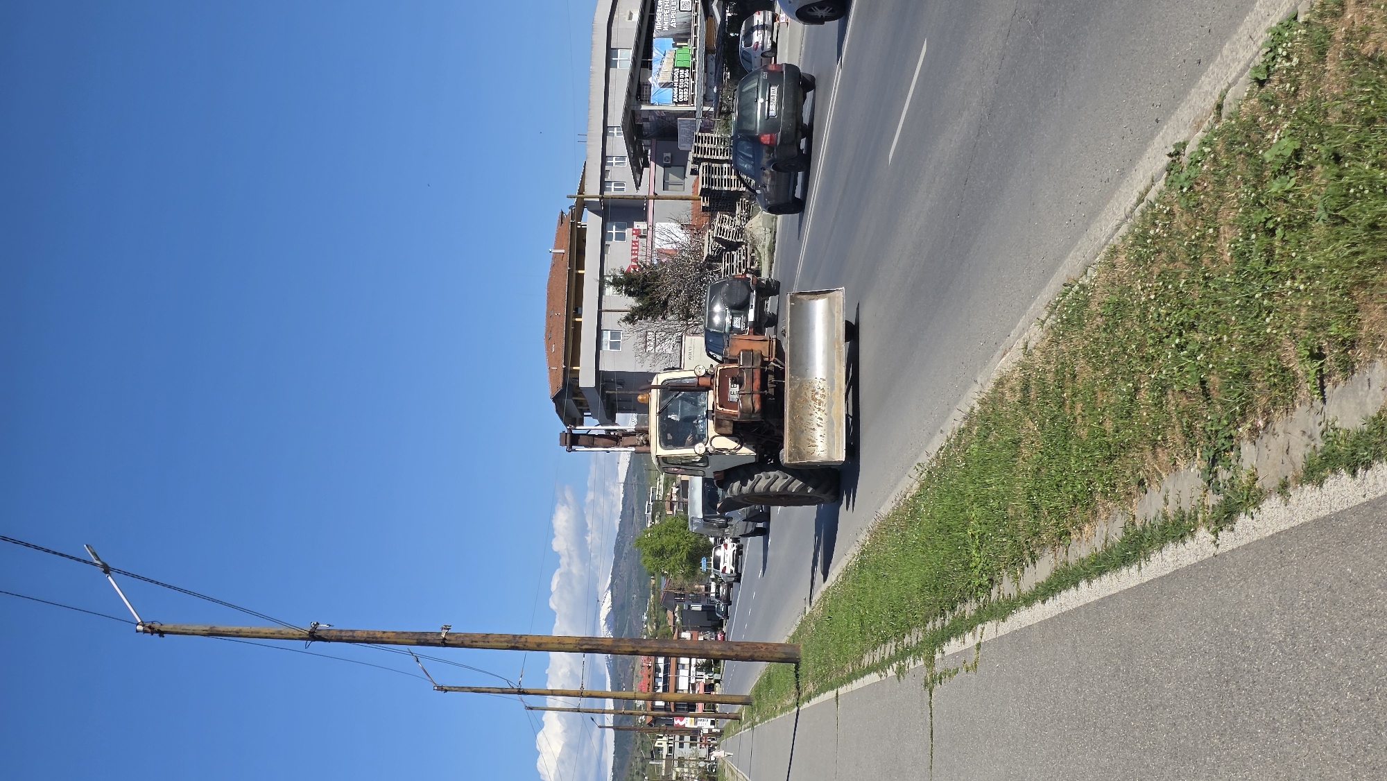 A long shot shows a street scene in Blagoevgrad, Bulgaria on a clear day. The image is rotated counterclockwise. A beige-colored tractor with a front-mounted blade travels on the right side of the road, facing the camera. Several cars are behind the tractor. Buildings are on the left side of the road, and grass grows along the curb on the right. Wooden utility poles line the left side of the road, against a bright blue sky. Mountains are visible in the distance.
