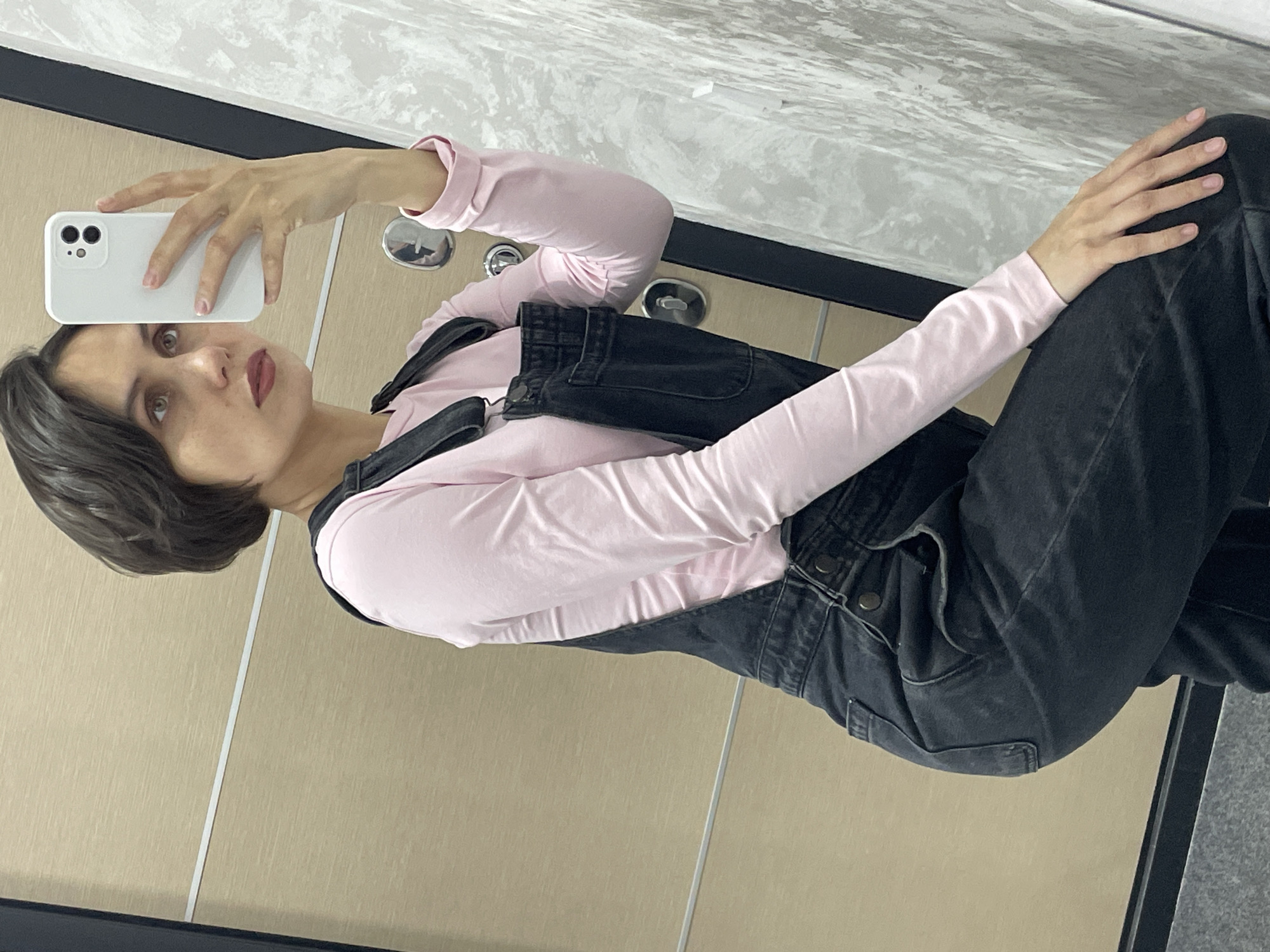 A young woman is captured in a mirror selfie taken indoors. She has short, dark hair and light-colored eyes, accentuated by dark red lipstick. She is wearing a light pink long-sleeved top layered under black denim overalls. Her left hand holds a white smartphone with dual rear cameras, pointing it towards her reflection to take the picture. Her visible fingernails are painted a light pink. Her right hand rests on her leg.
The background features a modern, clean aesthetic. Below the woman, a dark, flat surface or trim is visible. Above this, a light-colored, possibly wood-paneled or textured wall extends, with a thin white vertical line interrupting its surface. Further up, a lighter, textured or patterned wall, possibly resembling marble, completes the background. The lighting is bright and even, suggesting an indoor setting like a home or a contemporary building. The woman's expression is neutral, and she is looking directly into the camera/mirror. No visible text is present in the image. The original image is presented rotated on its side.