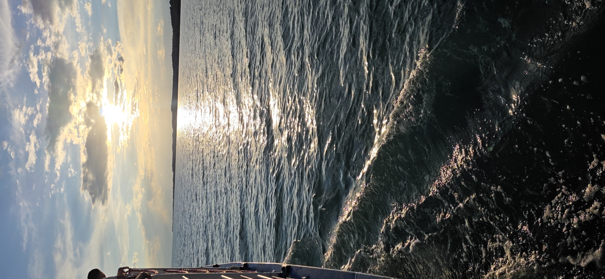 This is a vertical, medium shot photograph taken from the side of a vessel traveling across a body of water. The sun is setting, casting a golden sheen across the rippling surface of the water, which is dark blue in color. The sun is low in the sky, partially obscured by clouds, and is reflected on the water. The horizon line is visible in the distance, with a dark, undulating landmass on the far side of the water. The left side of the photograph shows the edge of the vessel, with a railing and some rope visible. The overall impression is one of tranquility and natural beauty. The location is the Issyk-Kul Lake, near the city of Pristan'-Przheval'sk, Kyrgyzstan.