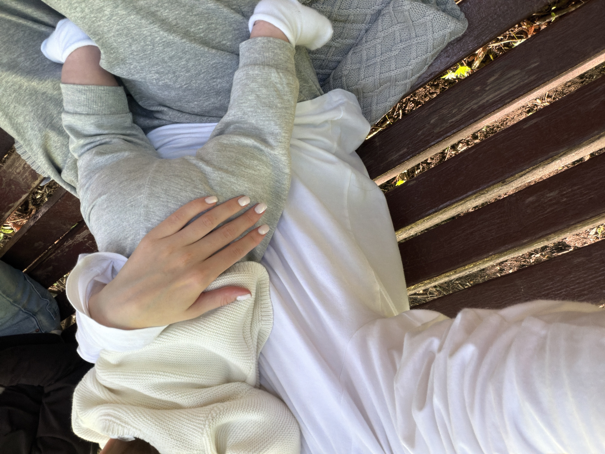 A top-down, close-up shot shows a person holding a baby on their lap while sitting on a wooden bench. The baby is wearing grey sweatpants and white socks. The person holding the baby has pale skin and white nail polish. They are wearing a white top and possibly a white sweater. The background shows a dark brown wooden bench with gaps between the planks and debris visible underneath. The setting appears to be outdoors, possibly a park. The location is identified as Khimki, Russia, although this is not visually confirmed.
