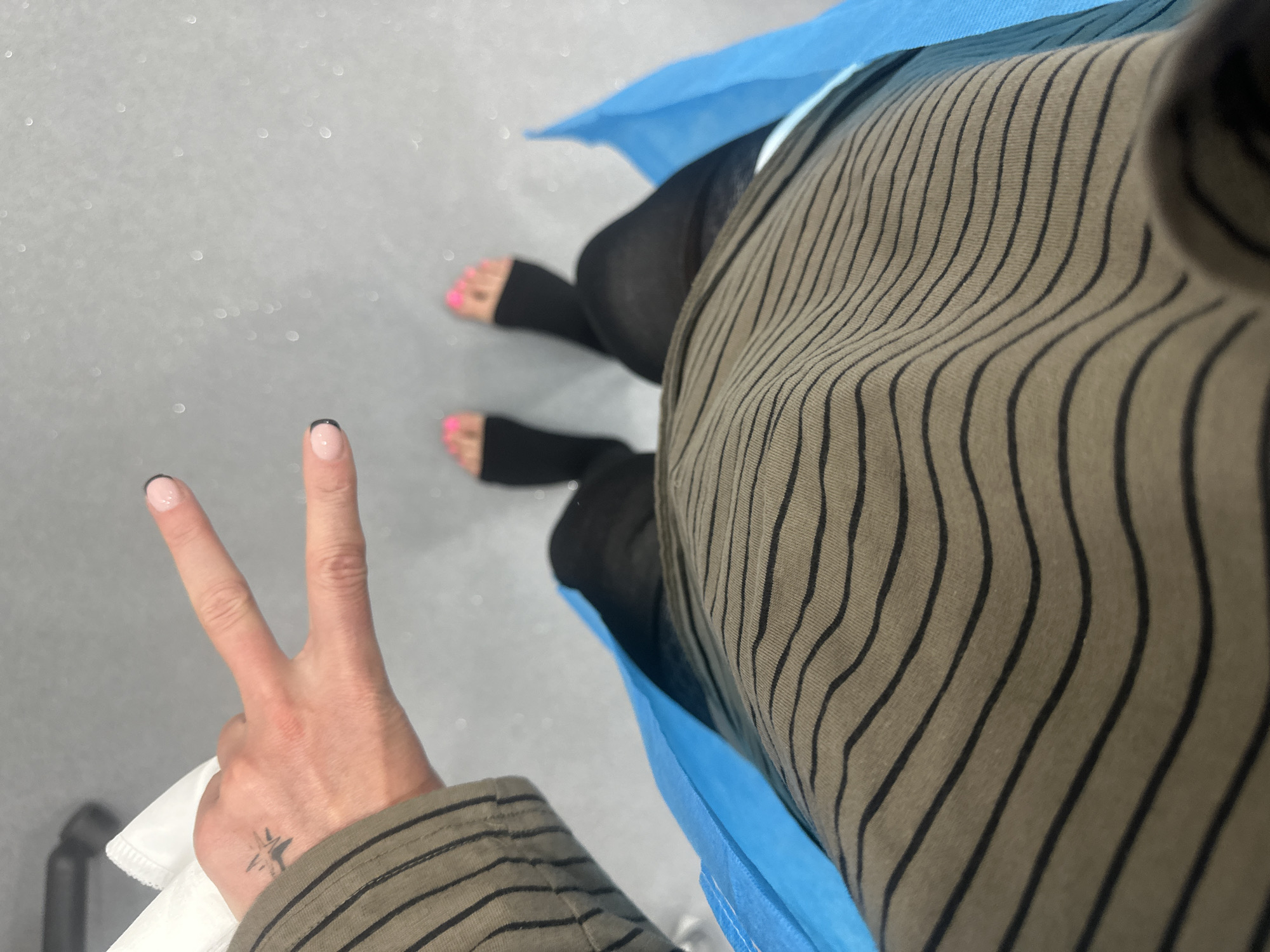 A downward-angle shot shows a person seated, wearing black leggings with foot openings and pink toenails. The person is wearing an olive green, pinstriped garment and is draped in a disposable blue gown. The left hand is raised in a peace sign, with nails painted nude with black tips. A small tattoo is visible on the hand. The floor is a light gray, speckled surface. The location is not identifiable as being in London.
