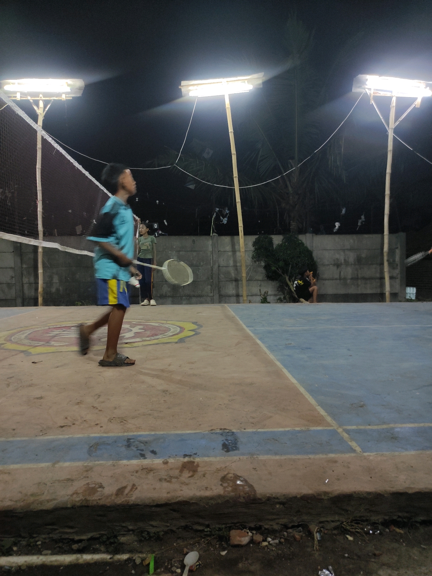 The image depicts an outdoor badminton court at night in Padalarang, Indonesia, illuminated by two bright floodlights mounted on bamboo poles. In the foreground, a young person wearing a blue t-shirt and yellow and blue shorts is in motion, holding a badminton racket and appearing to be playing. To the left, a badminton net is visible, stretched across the court. In the background, another young person in a green t-shirt stands near the net, also holding a racket. Further in the background, a concrete wall serves as a backdrop, and some trees and foliage are present. On the right side of the court, another individual is seated near a tree. The court surface is a mix of dirt and painted areas, with a blue line demarcating one section. The overall environment suggests a casual recreational setting.