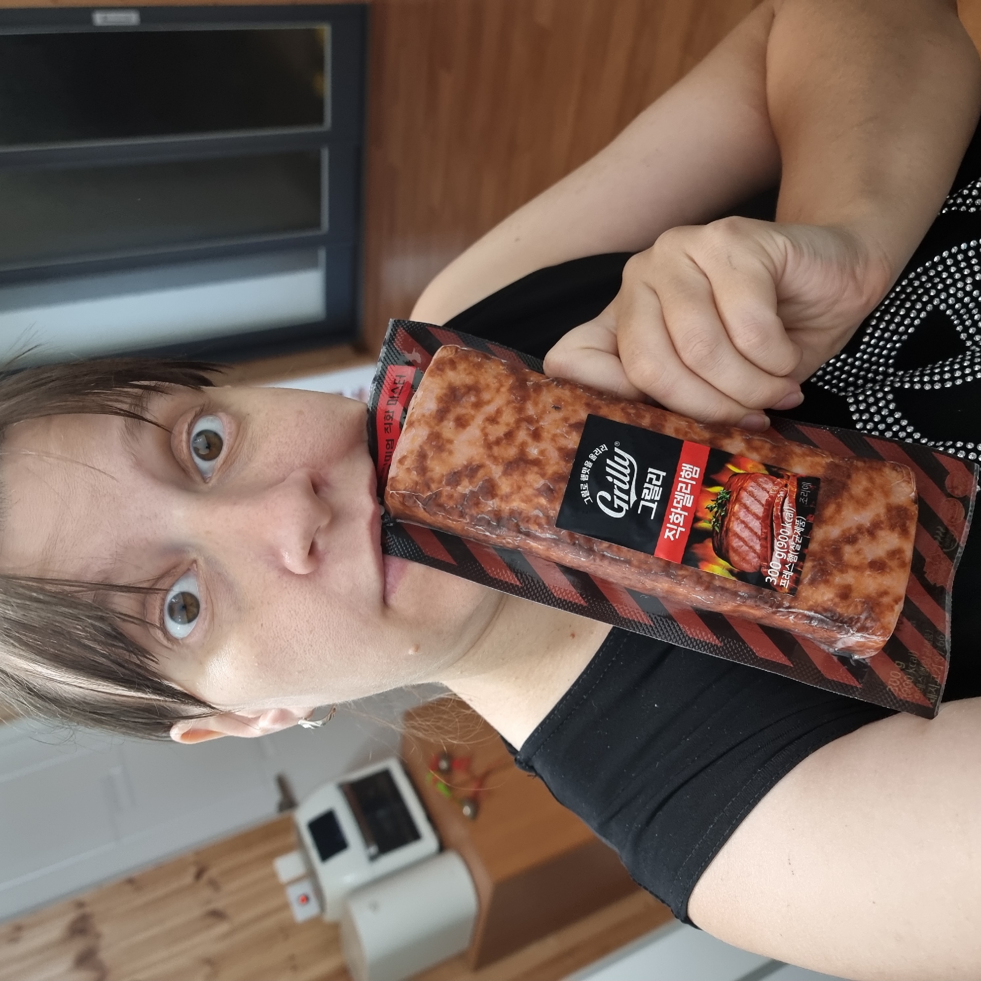 A medium shot shows a woman biting into a package of Grilly ham. She is wearing a black shirt with a silver stud pattern. The ham package is red and black with flames and images of grilled ham. In the background, a wooden wall is visible, along with a white appliance and a black window. The location appears to be indoors, possibly a kitchen or dining area.