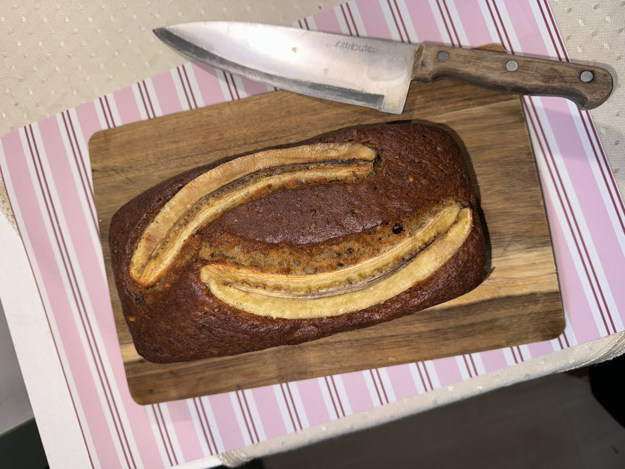 A high-angle shot shows a loaf of banana bread on a wooden cutting board, with a large knife lying on top. The banana bread is topped with sliced bananas. The cutting board sits on a pink and white striped placemat. The location is not discernible from the image. There are no people visible.
