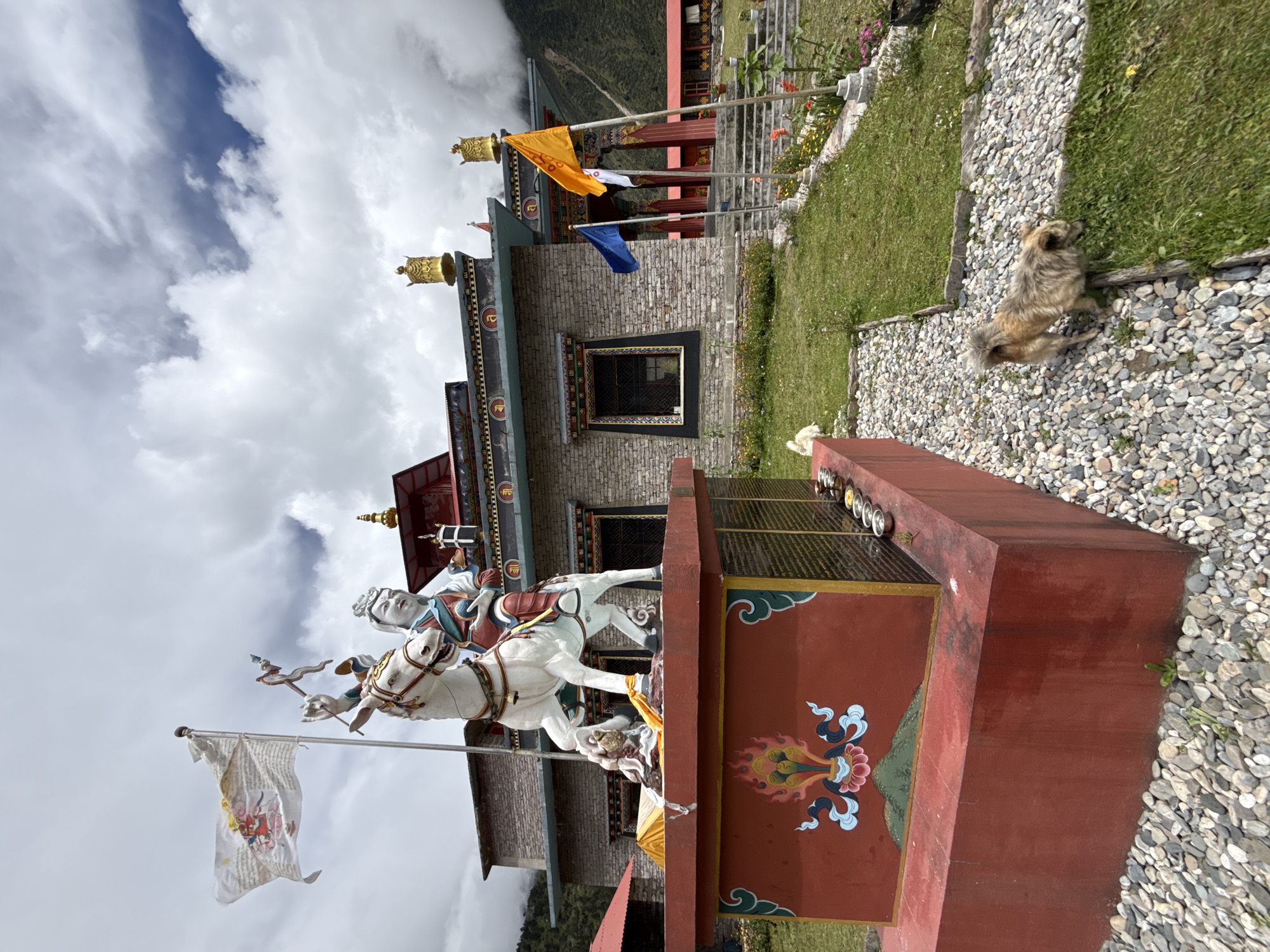 The image captures a serene scene at a monastery in Samagaon, Nepal, under a cloudy sky. In the foreground, a statue of a person on horseback stands prominently on a red pedestal adorned with colorful Buddhist motifs. To the right, a shaggy dog ambles across a gravel pathway leading to a grassy area dotted with flowers. Behind the statue, a traditional Buddhist building with brick walls and decorative golden elements on its roof is visible. Several flags, including a white one with intricate designs and colorful ones in orange and blue, flutter in the breeze. The overall environment suggests a peaceful and spiritual setting, typical of a monastery in the Nepalese Himalayas.