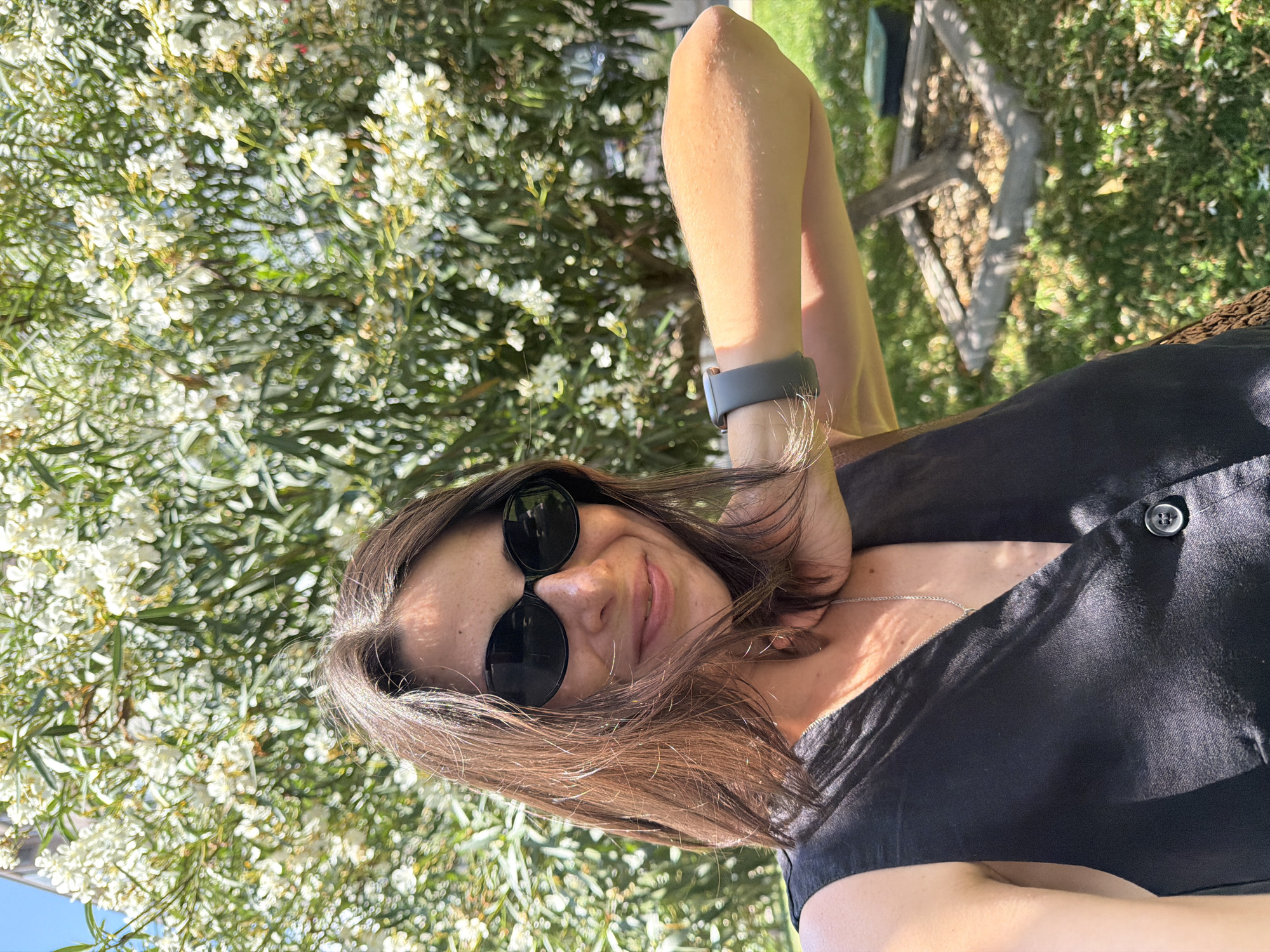 A woman is captured in a selfie outdoors, standing in front of a tree with white flowers in Podgorica, Montenegro. She is wearing a black sleeveless top, sunglasses, and a smartwatch. Her right arm is bent and resting on her neck. The background shows green grass and the white blossoms of the tree. The lighting suggests it is daytime.