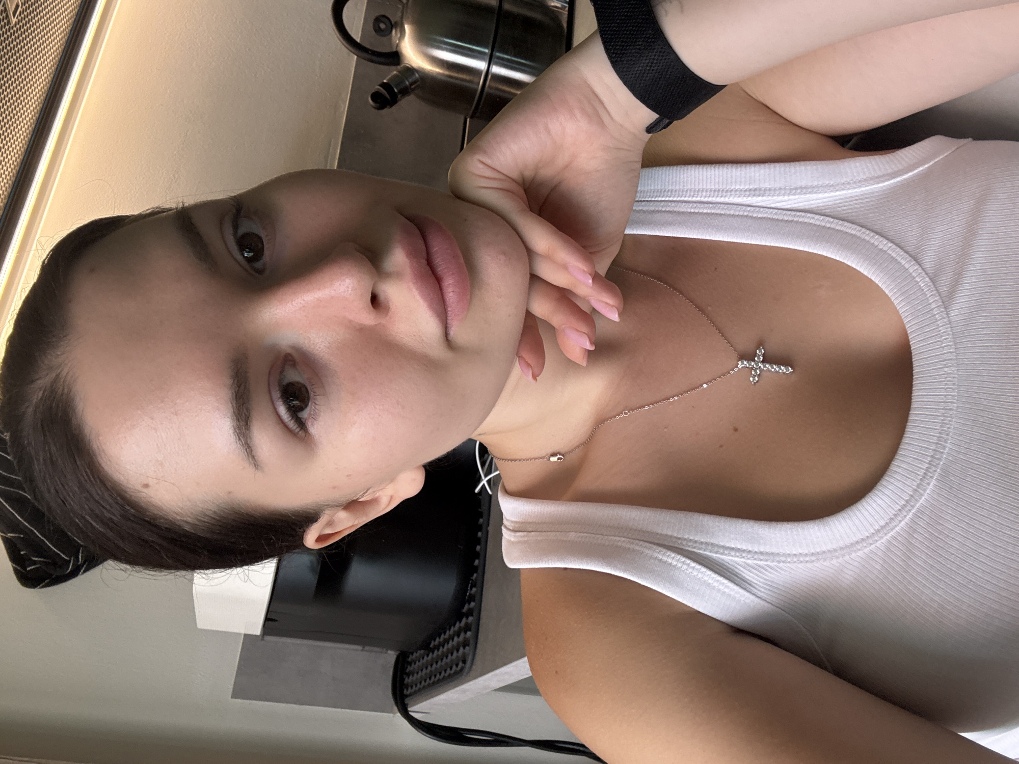 A young woman with dark hair pulled back is taking a selfie in what appears to be a kitchen. She is wearing a white ribbed tank top and a silver necklace with a cross pendant. Her left hand is resting on her chin. In the background, a black appliance and a stainless steel kettle are visible on a countertop. The lighting suggests it is daytime. There is no information available about the location being Prague, Czechia.