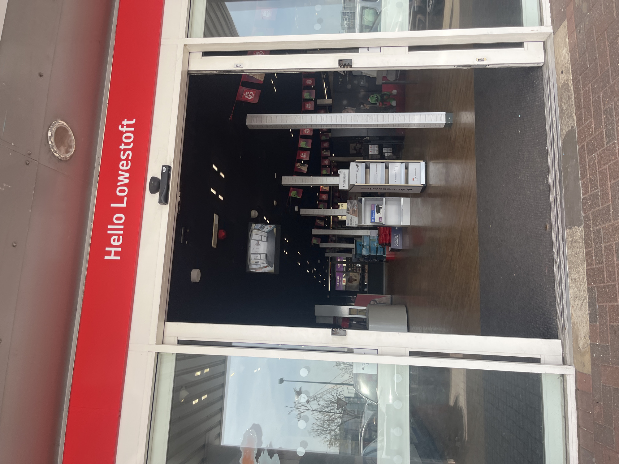 The image shows the entrance to a shop, identified by the sign "Hello Lowestoft" on the door frame, placing it in Lowestoft, East Suffolk, United Kingdom. The door is an automatic sliding glass door, currently open, revealing the shop's interior. The interior appears dimly lit, with displays of merchandise visible on shelves and hanging from the ceiling. The flooring transitions from wood-like planks to a darker, possibly carpeted area further inside. The exterior includes a brick walkway and a glimpse of the outside world through the glass doors, showing a cloudy sky and trees. There are no people visible in the image.