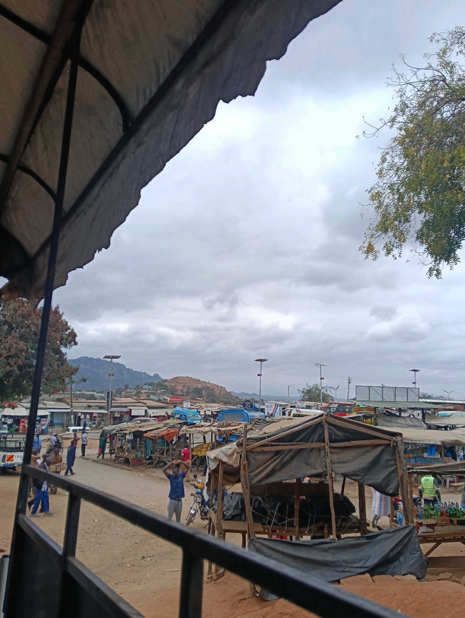 The image depicts a bustling outdoor market scene in Kivou Ward, Kenya, under a cloudy sky. The foreground is dominated by the weathered, dark fabric and metal structure of a canopy, suggesting the perspective of someone observing from a vehicle or covered stall. In the midground, a dirt road winds through a collection of market stalls, many with rustic wooden frames and coverings made of corrugated metal or fabric.
Merchandise is displayed at the stalls, including bottles of beverages and what appear to be food items. People are actively engaged in market activities: some are walking along the road, others are interacting at the stalls, and a few are operating motorbikes and a small truck. A man in a blue shirt and grey pants stands with his hands raised in the air in the center of the scene, while another individual in a high-visibility vest is visible at a stall further to the right, attending to bottles.
In the background, more market stalls and buildings are visible, extending towards a hilly landscape. Several vehicles, including buses and trucks, are parked or moving through the market area. The overall atmosphere is one of everyday commerce and community activity in a semi-urban or rural Kenyan setting. The weather appears overcast, with a diffused light filtering through the clouds.