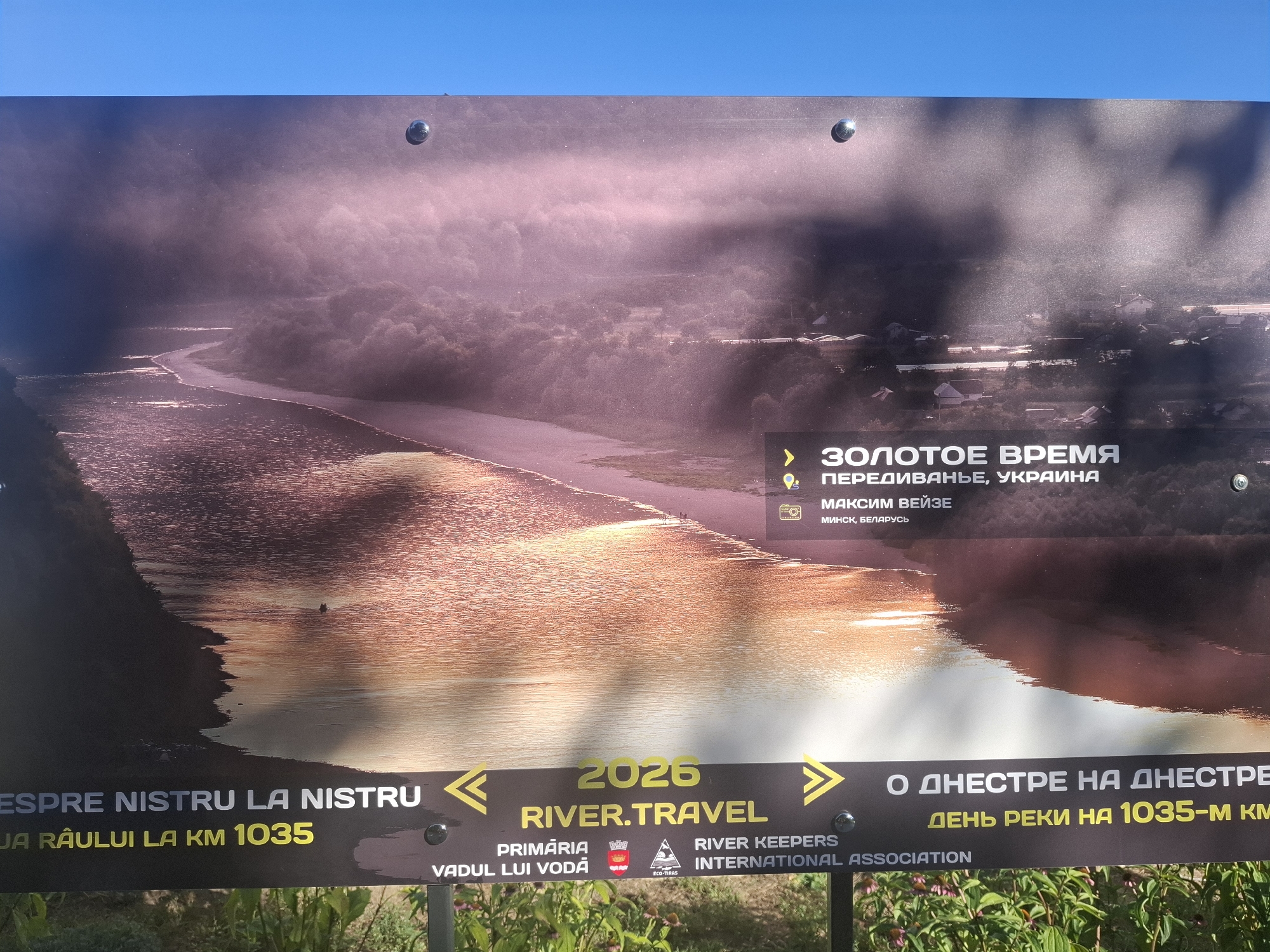 The image shows a large outdoor sign with a landscape photograph of a river winding through a valley, bathed in hazy sunlight. The sky above is a clear, bright blue. Text overlays the photograph and a dark band at the bottom of the sign.
In the upper right quadrant of the sign, there is text in Russian that reads "ЗОЛОТОЕ ВРЕМЯ" (Golden Time) followed by "ПЕРЕДИВАНЬЕ, УКРАИНА" (Peredivane, Ukraine) and "МАКСИМ ВЕЙЗЕ МИНСК, БЕЛАРУСЬ" (Maxim Veize, Minsk, Belarus).
The dark band at the bottom of the sign contains text in Romanian and Russian. On the left, in Romanian, it says "ESPRE NISTRU LA NISTRU" (Towards the Nistru at the Nistru) and "UA RÂULUI LA KM 1035" (of the River at KM 1035). In the center, in yellow text, is "2026 RIVER.TRAVEL". Below this, in Romanian, is "PRIMĂRIA VADUL LUI VODĂ" (City Hall Vadul lui Voda) with a coat of arms to its right. To the right of "RIVER.TRAVEL", in Russian, it reads "О ДНЕСТРЕ НА ДНЕСТРЕ" (About the Dniester on the Dniester) and "ДЕНЬ РЕКИ НА 1035-М КМ" (River Day at 1035-th KM). Further to the right is text in English that appears to be "RIVER KEEPERS INTERNATIONAL ASSOCIATION".
The photograph itself depicts a wide, calm river reflecting the golden light of the sun, with lush green trees lining its banks and a scattering of buildings in the distance. A small, dark boat or object is visible on the river in the middle distance.
The sign appears to be mounted outdoors, possibly as part of an informational display or advertisement related to river tourism or an event. The presence of vegetation and flowers in the foreground suggests the sign is situated in a public outdoor space. The location mentioned on the sign, Vadul lui Voda, is a town in Moldova known for its natural beauty and recreational areas along the Dniester River.