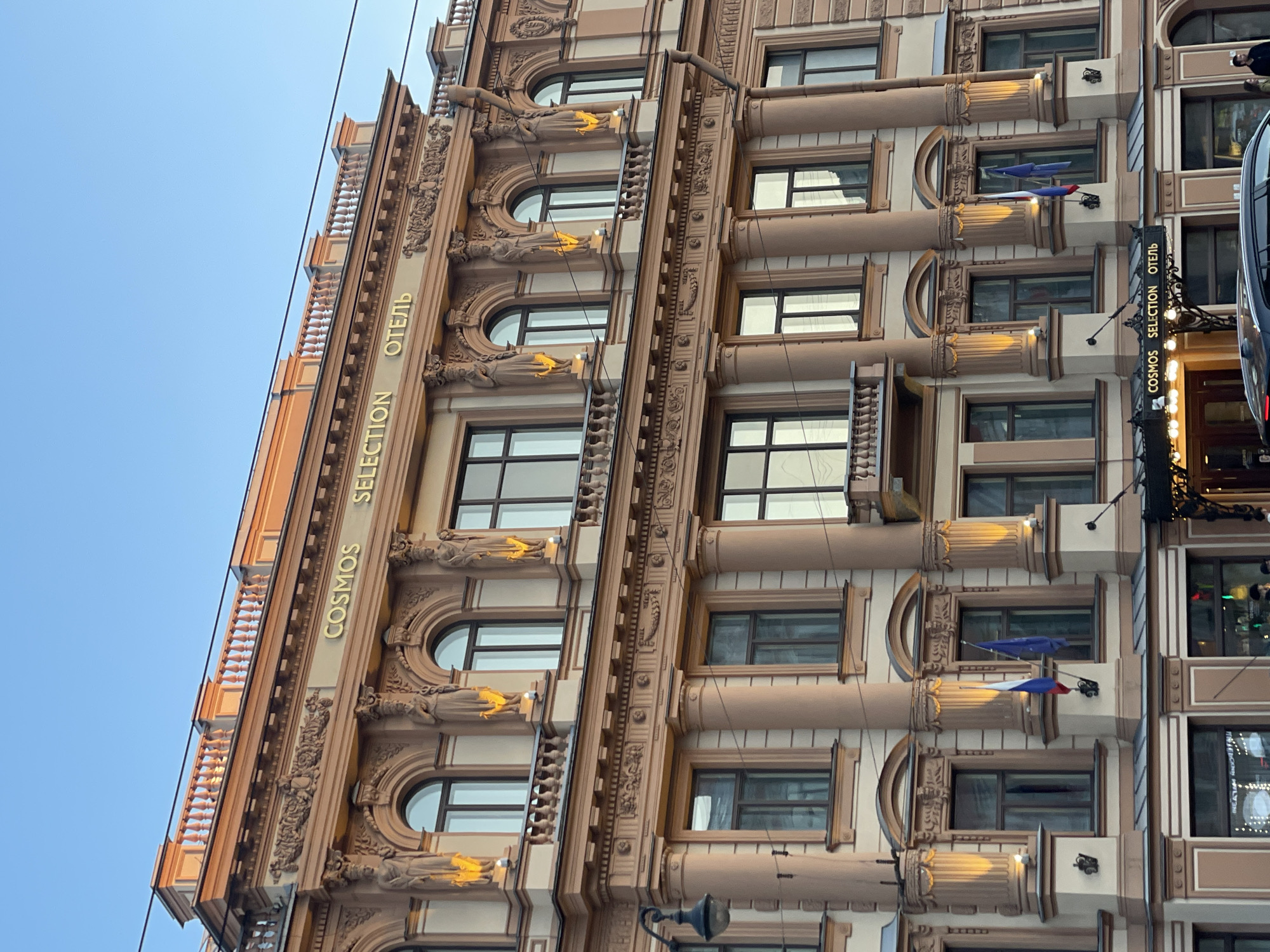 The image shows a section of the Cosmos Selection Hotel in Saint Petersburg, Russia. The building has an ornate facade with architectural details such as statues, arched windows, and columns. The name "COSMOS SELECTION ОТЕЛЬ" (hotel) is written vertically in gold lettering on the building's exterior. French flags are mounted on the building. A street lamp is visible on the lower right, and a section of a vehicle is visible at the upper right. The sky is clear and blue.