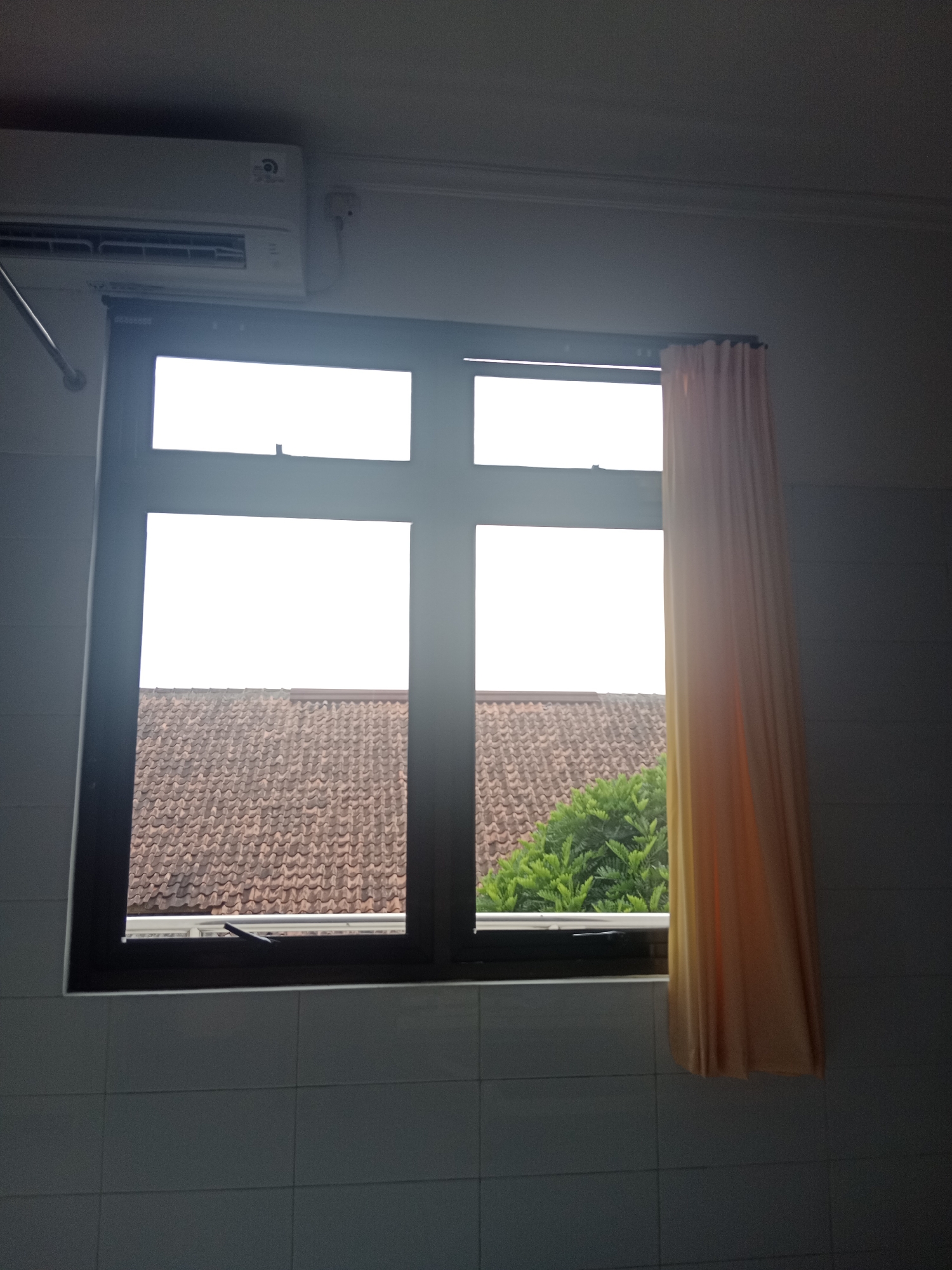 A dimly lit indoor scene captures a window with a dark frame, divided into four panes by a central crossbar. The top two panes are overexposed, showing a bright white sky. The bottom two panes reveal a view of terracotta roof tiles and lush green trees. To the right of the window, a pale orange curtain is partially drawn, obscuring the right side of the view. Above the window, a white air conditioner unit is mounted on a light grey wall. The lower portion of the wall is tiled with square, light grey tiles. The scene appears to be from an interior in Yogyakarta, Indonesia.