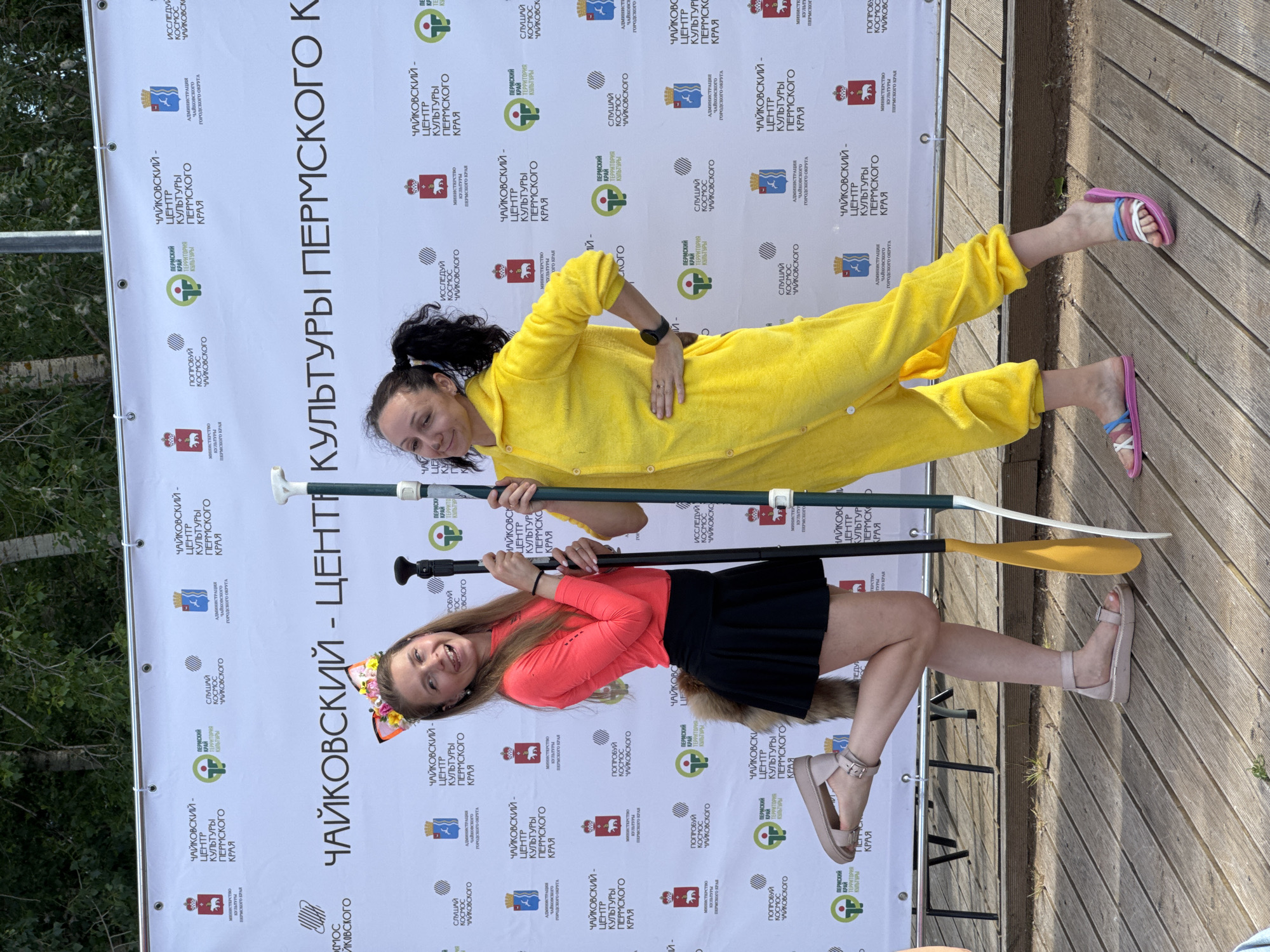 Two women are posing for a photo in front of a banner with logos. The woman on the left is wearing a pink long-sleeved shirt, a black pleated skirt, and cat ears with flowers. She is also wearing tan sandals with ankle straps and has a furry tail attached to her skirt. The woman on the right is wearing a yellow fuzzy onesie with a hood. Both women are holding upright poles, which appear to be part of paddles or oars. The banner behind them features various logos, including what appears to be the coat of arms of the Perm Krai region and text in Russian that translates to "Chaykovsky - Center of Culture of Perm Krai." The setting seems to be outdoors on a wooden platform or deck, with trees visible in the background. The lighting suggests it is daytime. This event is likely related to a cultural festival or initiative in Chaykovsky, Russia.