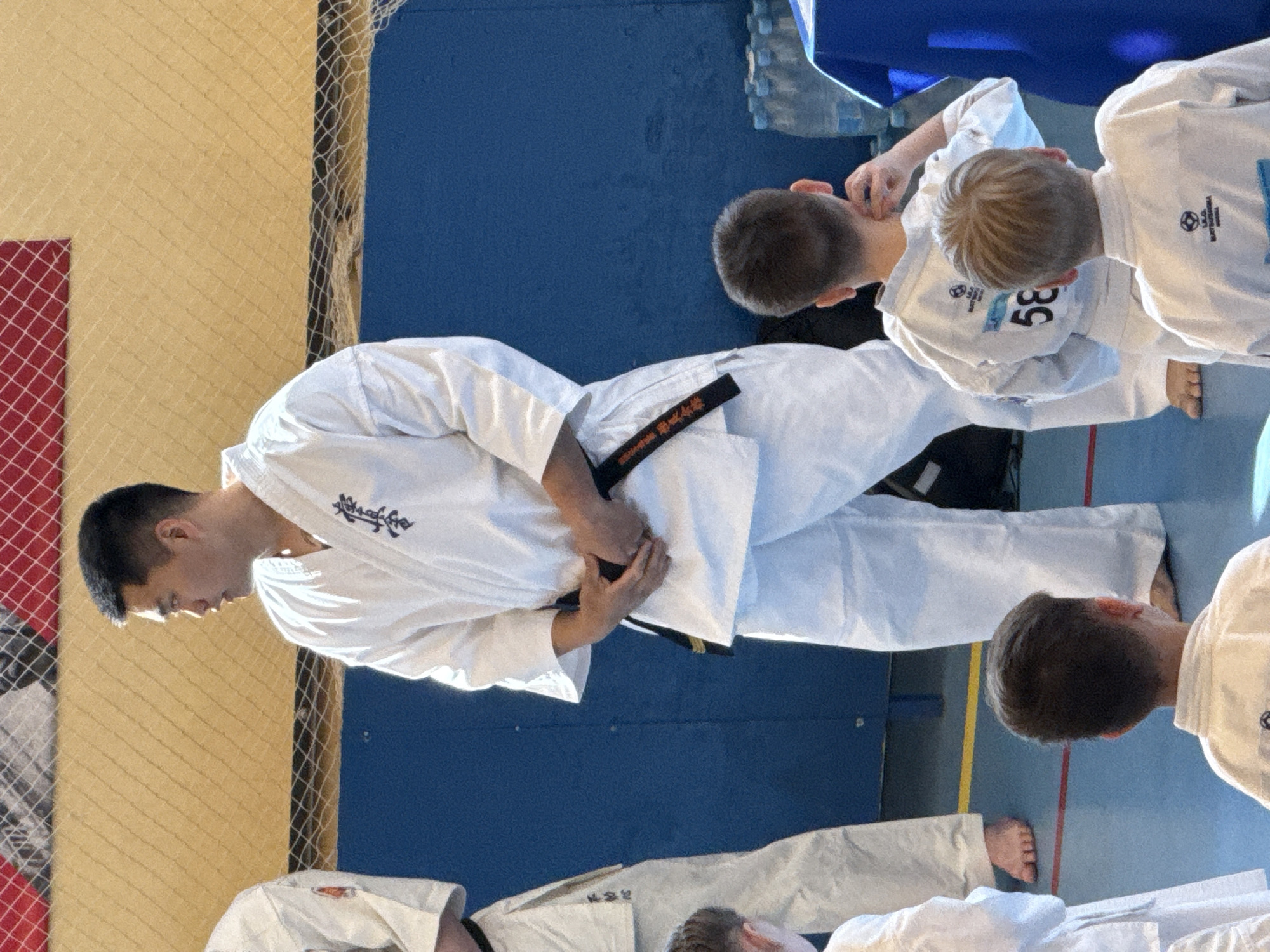 The image depicts a man wearing a white karate uniform and a black belt, surrounded by younger individuals, also in karate uniforms. They appear to be in a gym setting. The man seems to be adjusting his belt. A net and patterned wall are visible behind him. Other objects include water bottles and a table. While Yekaterinburg, Russia, may be the location, there is no discernible feature to indicate the location or place, therefore location is not discernible.