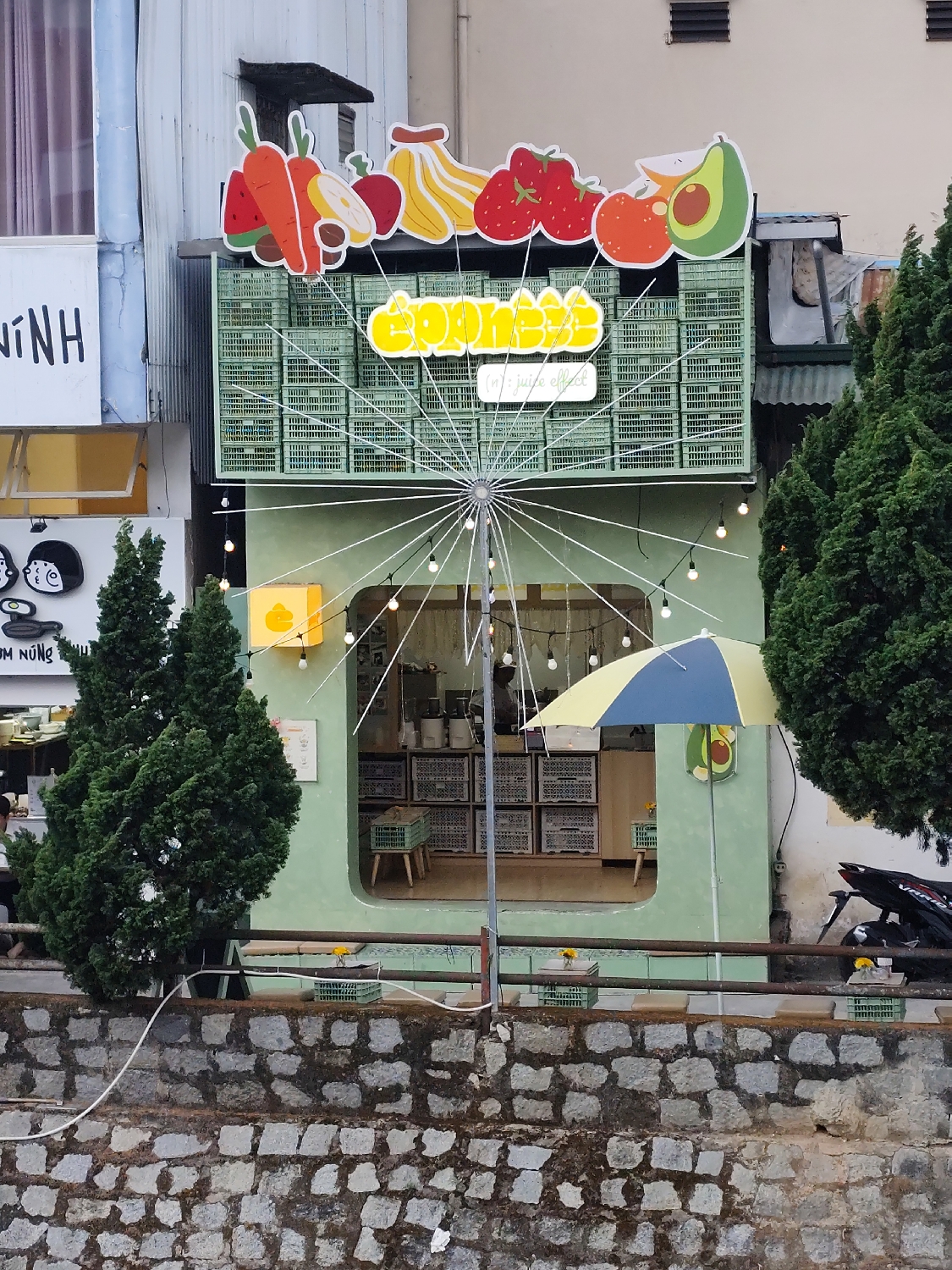 A vibrant and creatively designed juice bar, named "Coobeez," stands out in an urban or semi-urban setting, likely in Da Lat, Vietnam, as indicated by the location context and visible Vietnamese text. The two-story establishment features a light green facade, exuding a cheerful and inviting ambiance.
The upper part of the shop's front is adorned with large, colorful cutouts of various fruits, including watermelon, carrots, lemon, bananas, strawberries, an orange-like fruit, and an avocado, hinting at its fresh beverage offerings. Below these, the shop's name "Coobeez" is displayed in bright yellow, with "in' juice effect" written beneath in smaller letters. The second story's front is uniquely decorated with numerous light green plastic crates, arranged in a grid pattern to create a textured and distinctive appearance.
Through a large, open archway on the ground floor, the interior of the shop is visible, showing more green crates ingeniously used as shelving or part of the counter, along with small stools crafted from similar crates. A yellow and blue umbrella is positioned inside, suggesting a casual, open-air setup. Strings of warm white light bulbs are hung decoratively around the entrance and leading down from a central pole, giving the space a cozy, illuminated glow, suggesting it's dusk or evening.
In the foreground, a sturdy stone retaining wall separates the shop from the viewer, with small green crates holding yellow flowers placed along its top edge as simple decorations. Two slender, conical evergreen trees flank the left side of the shop, while a larger, dense tree partially obscures the right side, where a black motorcycle is parked. Adjacent buildings are partially visible; a plain white structure to the left bears the Vietnamese text "DẠ NINH," and a black sign below it reads "TÚM NÚNG." The overall impression is one of a whimsical and welcoming local business.