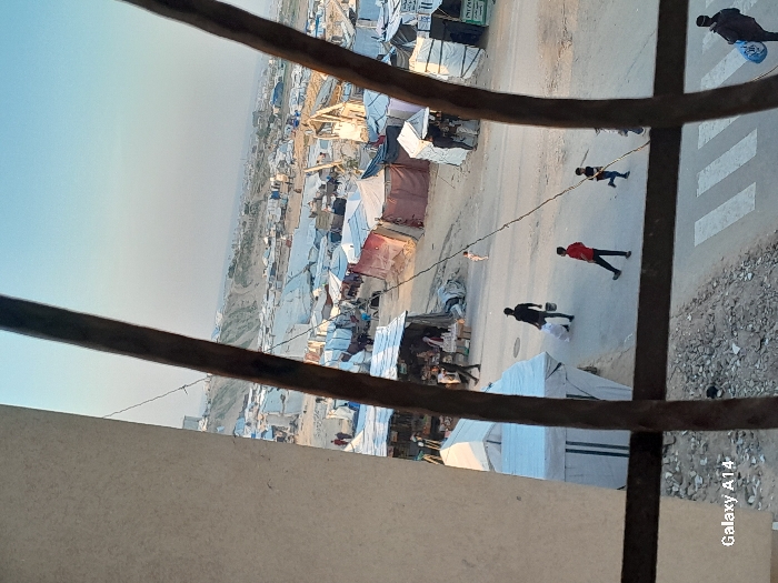 The image, framed by two dark, irregular vertical bars on the right and a light vertical surface on the left, depicts a sprawling informal settlement in Khan Yunis, Palestinian Territory, under a clear, pale blue sky. Numerous makeshift shelters and tents, predominantly white, grey, and some reddish-brown, are densely packed across uneven terrain. A dirt road, with a paved section featuring a white crosswalk in the mid-ground, traverses the scene.
Multiple individuals are visible on or near the road:
- In the lower right, a person in dark attire walks right-to-left, carrying a blue bag on their left shoulder and a blue object in their right hand.
- Above the crosswalk, a child in a dark shirt and light trousers runs right-to-left, holding a stick.
- To the left of the child, a person in a red shirt and dark trousers walks left-to-right.
- Further left, an individual in dark clothing walks right-to-left, carrying a white bag over their shoulder and a white bucket in their left hand.
- Near the center, a small, light-colored animal, possibly a dog, walks rightward.
- Several other individuals are seen walking amidst the structures in the mid-ground, some near what appear to be market stalls.
The settlement extends into the distance, with a large, barren, light-colored hill or elevated area forming the background horizon. Rocky debris is visible along the left edge of the road.