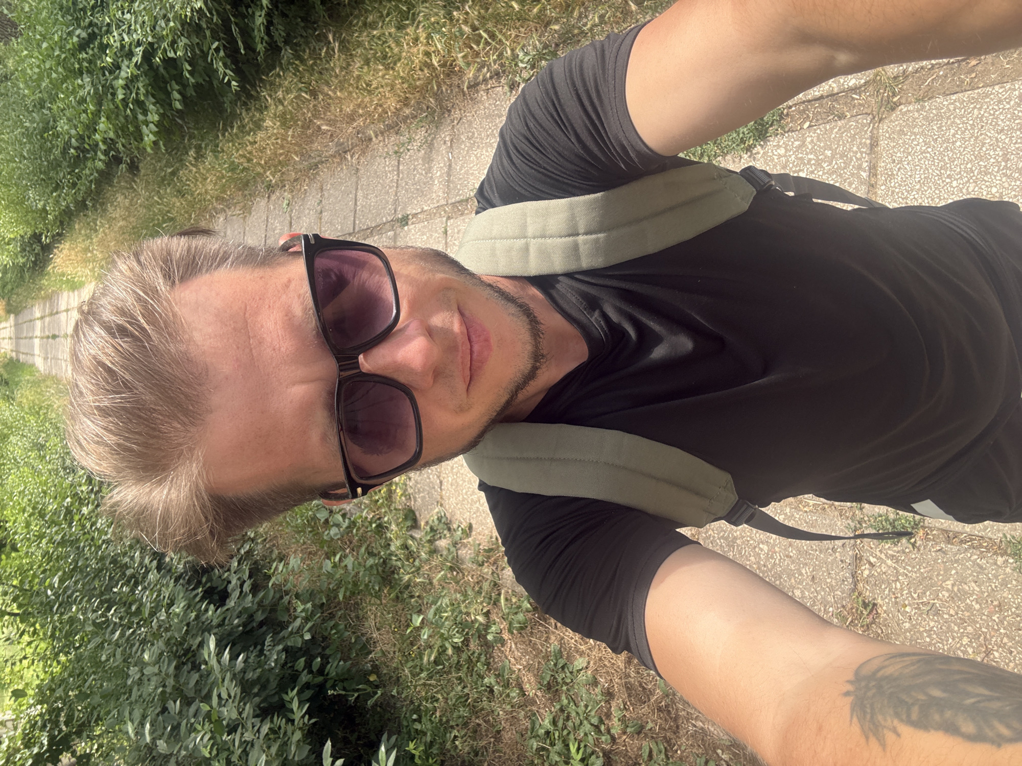 The image is a selfie taken by a man with blond hair and sunglasses, wearing a black t-shirt and a light green backpack. He is standing on a paved path in a park or natural area. The path is lined with dense green foliage on the left and drier grass and some foliage on the right. The path recedes into the distance, suggesting a linear walkway. The lighting indicates it is daytime, likely sunny, with shadows visible on the path. The man's left arm, tattooed with a feather design, is extended towards the camera. The background is slightly blurred, focusing attention on the man. The overall scene suggests an outdoor activity, possibly hiking or walking. The location is identified as Aeroflotsky, Ukraine, though this cannot be visually confirmed from the image itself.