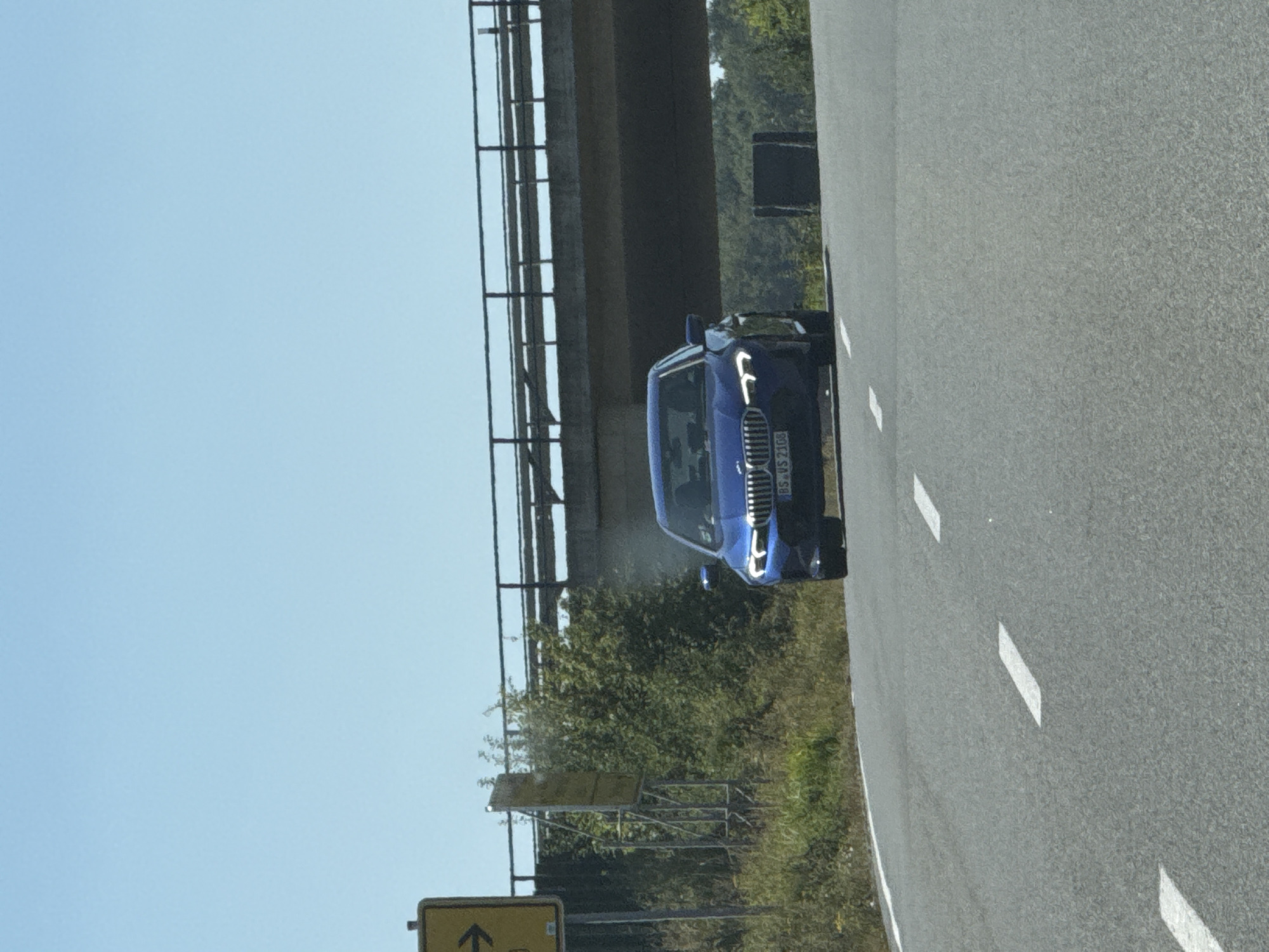 A blue BMW sedan, license plate BS VS 2108, drives towards the camera on a two-lane asphalt road. The car is positioned in the center of the right lane. The road has white dashed lane markings. In the background, a concrete overpass with a metal railing spans across the road. To the left of the road, there is a yellow road sign with a black arrow pointing right and a smaller, faded yellow sign attached to a metal frame. Green bushes and trees are visible on the left and right sides of the road, with denser foliage and trees behind the overpass. The sky is clear and blue. The image appears to have been taken on a sunny day. The scene is likely a rural or semi-rural area in or near Bad Sachsa, Germany, given the presence of German license plates and road signs.