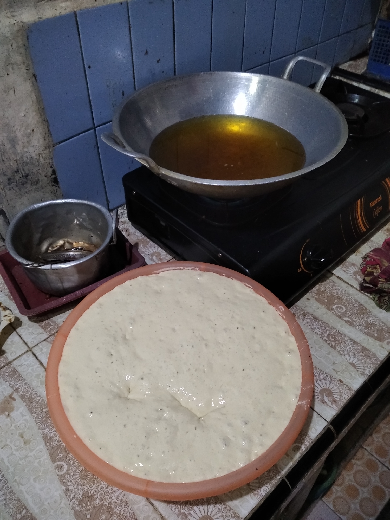 A close-up, overhead shot shows a cooking scene on a tiled surface in what appears to be an outdoor or semi-outdoor kitchen. In the foreground, a large, pale, unrisen dough is spread in a wide, shallow orange bowl, its surface uneven with visible air pockets. To the right and behind the dough, a black gas stove with a silver wok filled with hot, amber-colored oil sits on one of its burners. The wok has two handles. To the left of the wok, a smaller metal pot with a strainer inside rests on a red tray, the pot containing some dark residue. The background features blue tiled walls with some grime and a glimpse of a blue plastic grate on the far right. The overall impression is of food preparation in a humble, possibly rustic, kitchen setting. This scene is not distinguishable as being in Rambipuji, Indonesia without additional context.