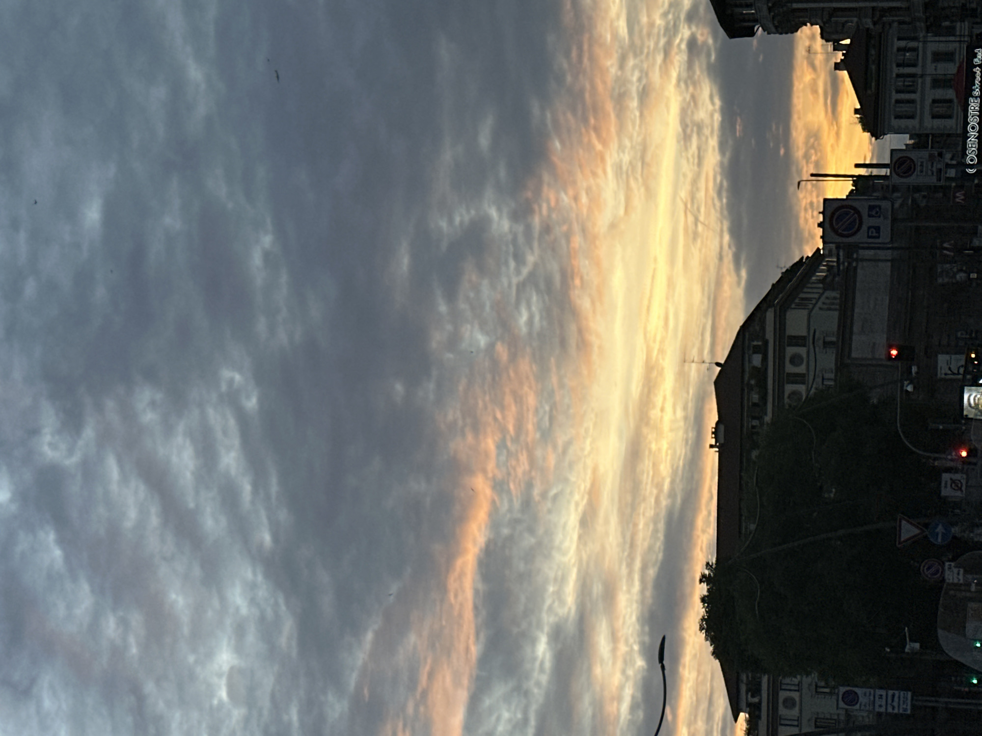 The image shows a sunset over a street in Milan, Italy. The sky is filled with clouds that are illuminated by the setting sun, casting shades of gray, white, and orange. Below the sky, the silhouettes of buildings and trees line the street. Streetlights and traffic signs are visible. The street appears relatively quiet, with no visible people or cars at the immediate moment of the capture. A sign reading "CO SENOSTRE Street Food" is visible on one of the buildings.