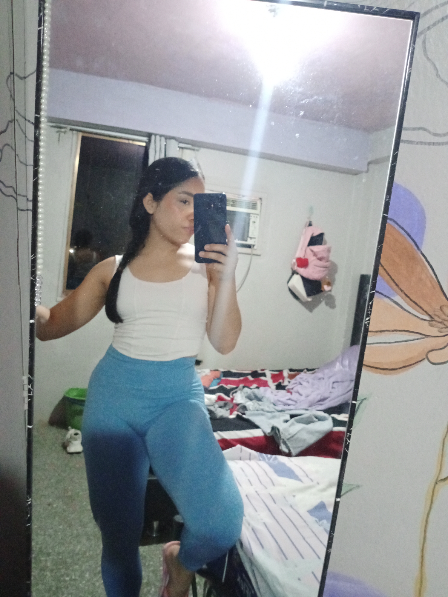 A young woman stands in front of a mirror, taking a selfie with a dark-colored smartphone. She is wearing a white top and blue leggings. Her hair is pulled back into a braid.
Behind her, the room is modestly furnished. There is a bed with rumpled bedding, a green basket on the floor, and personal items like a backpack hanging on the wall. An air conditioning unit is mounted on the wall above the bed.
The location is likely indoors, possibly a bedroom in a private residence. I cannot confirm the location in the city of Valle de La Pascua, Venezuela, from this image alone.