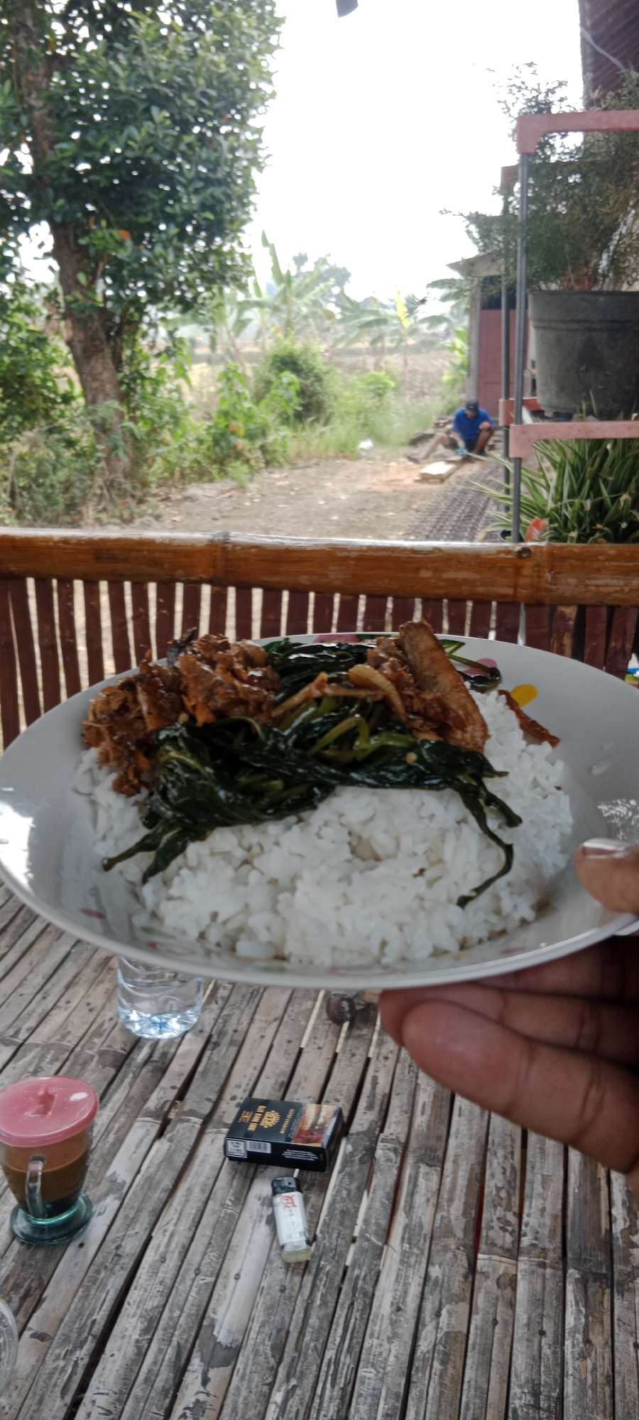 A close-up, eye-level shot shows a plate of rice topped with vegetables and meat, held by a person's hand. The plate is set on a bamboo table in an outdoor environment. In the background, a person is visible working in what appears to be a yard or garden. The overall setting suggests a casual meal in a rural or semi-rural area, possibly in Pelang, Indonesia, given the distinct bamboo construction and vegetation. A glass of iced coffee, a pack of cigarettes, and a lighter are also present on the table.