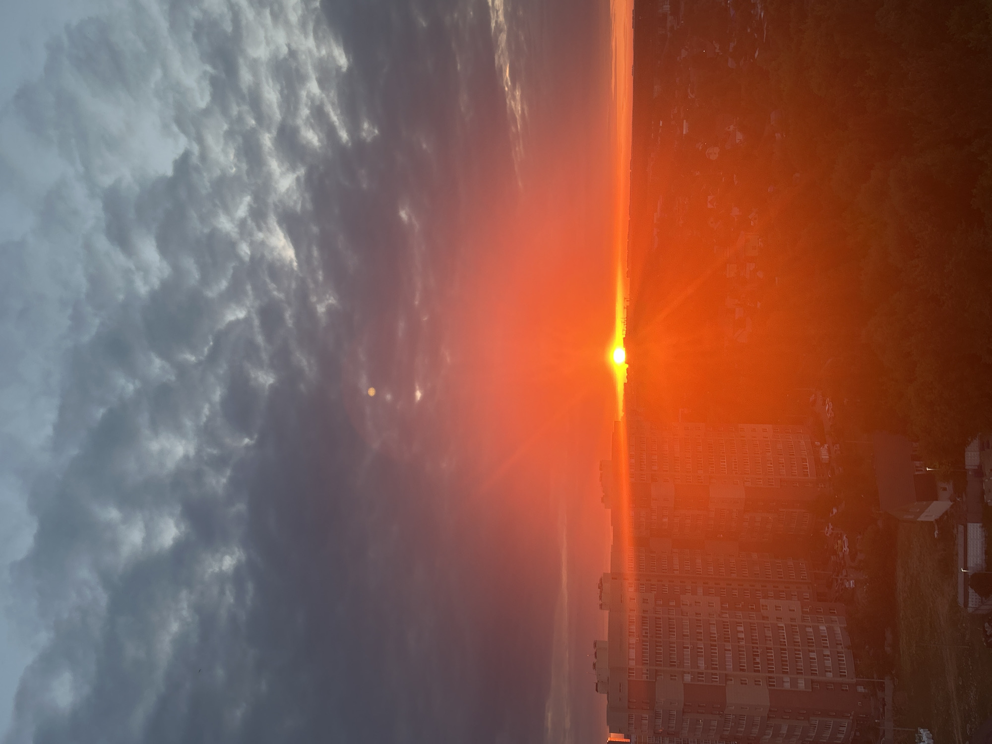 This image depicts a sunset over the city of Ivanteyevka, Russia. The sky is filled with dramatic clouds, ranging from dark gray to bright orange and red hues, indicating the sun is low on the horizon. The sun itself is a brilliant, glowing orb, casting a warm, intense light across the scene. In the foreground, several multi-story apartment buildings are visible, their windows reflecting the sunset's light. Some trees and smaller structures can be seen in the distance, interspersed with hints of more urban development. The overall atmosphere is one of a striking and vibrant natural phenomenon occurring over a populated area.