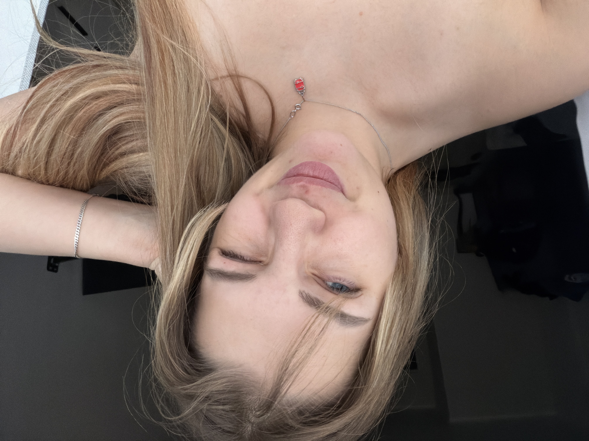 A woman is lying down with her head tilted. She has blonde hair, fair skin, and one eye closed. She is wearing a silver necklace with a red pendant. Her right arm is bent and positioned near her head. She is also wearing a silver bracelet on her right wrist. The background is a mix of light and dark surfaces. The image does not provide enough information to determine the location in Kazan, Russia.