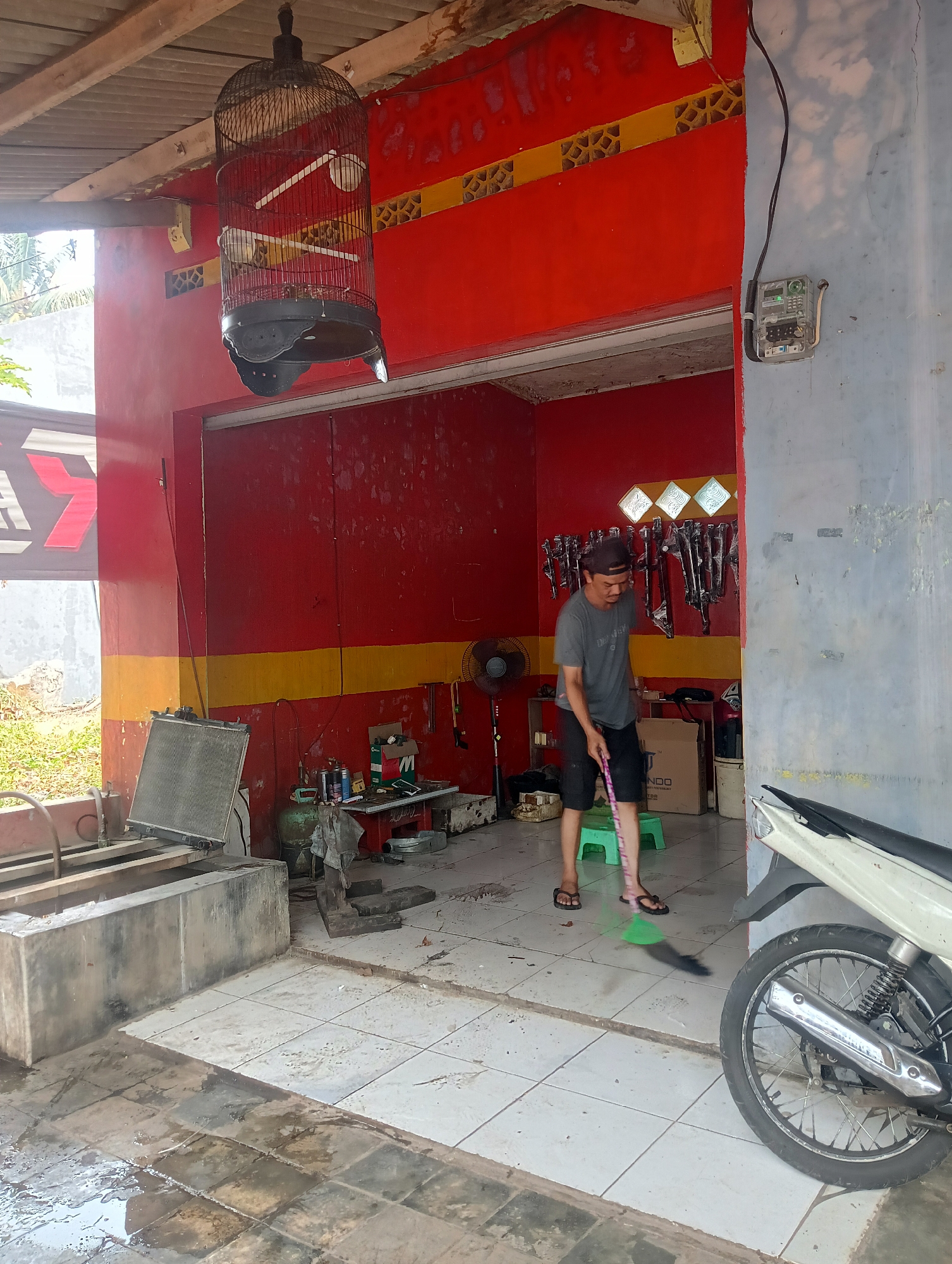 A man is sweeping the floor of what appears to be a motorcycle repair shop or similar establishment in Manggari, Indonesia. The shop has red walls with a yellow stripe and a corrugated metal roof. A bird cage hangs from the ceiling, containing a bird. In the foreground, a motorcycle is parked. Tools and equipment, including a radiator, are visible in the background. The floor is tiled and wet in some areas. The overall impression is of a functional, working space.