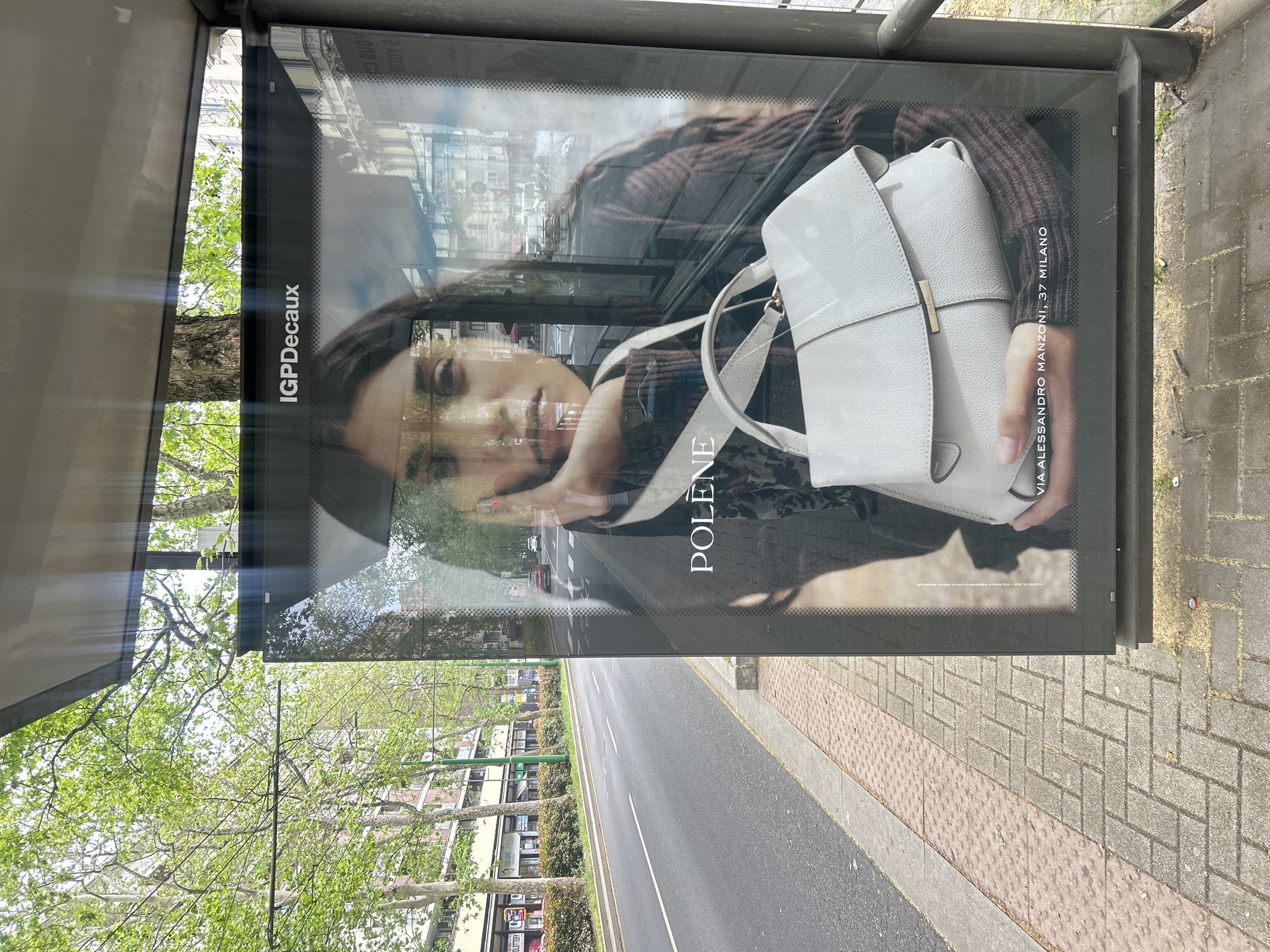 The image shows a bus stop in Milan, Italy. Inside the bus stop is an advertisement for Polene, a handbag brand. The ad features a woman holding a white handbag. The address "VIA ALESSANDRO MANZONI, 37 MILANO" is printed on the advertisement. The bus stop is located next to a road with trees lining the sidewalk. The sidewalk is paved with brick and stone. Buildings can be seen across the street.