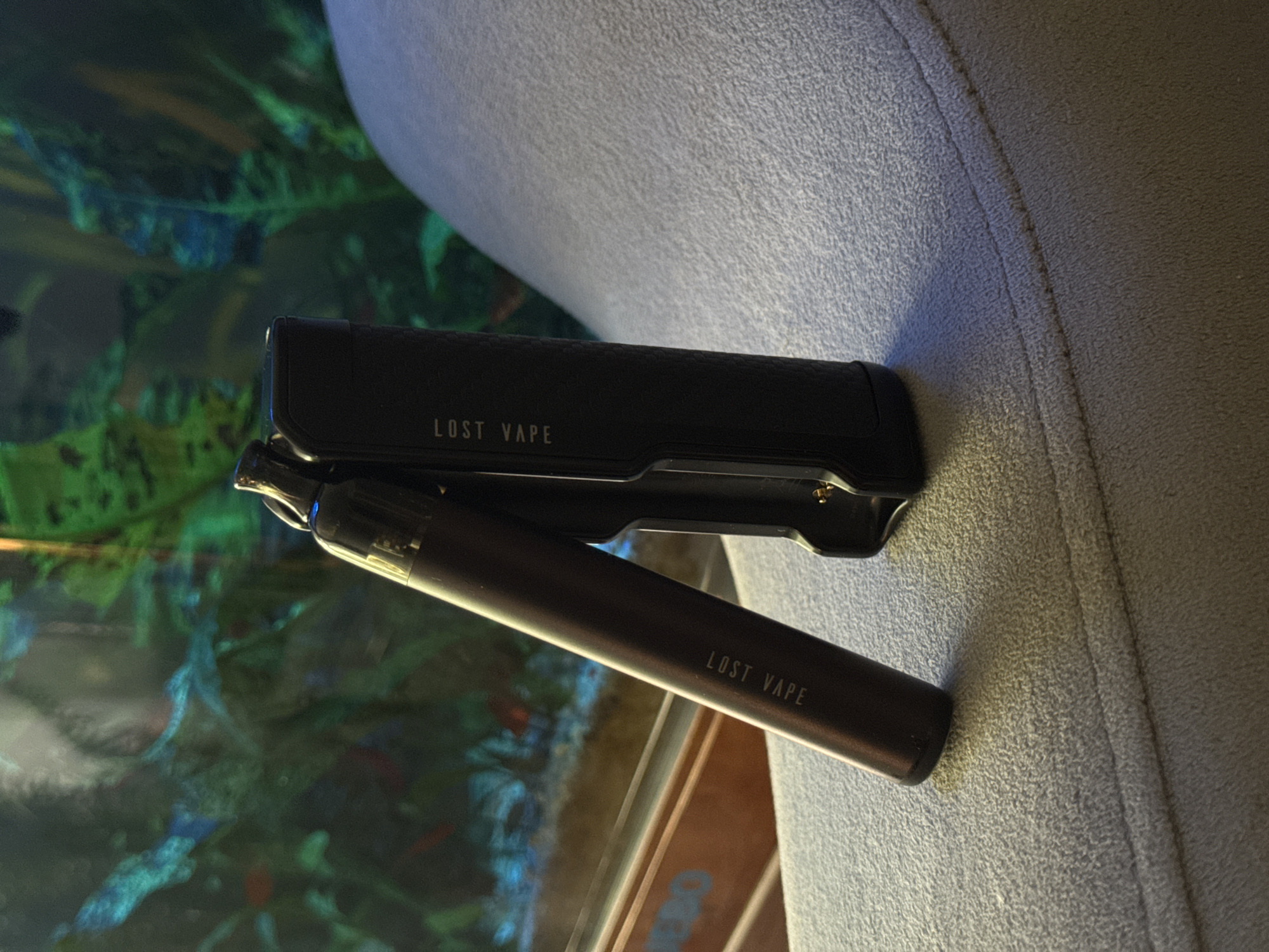 The image displays two vaping devices resting on a textured, light-colored surface, possibly upholstery or fabric. The device on the left is a dark brown cylindrical pod system with "LOST VAPE" printed vertically in white lettering. The device on the right is a black, rectangular box mod with "LOST VAPE" also printed vertically in white lettering. Behind the devices, a blurred background suggests an aquarium with green aquatic plants and possibly fish, indicated by hints of red and blue in the water. The lighting appears to be artificial, casting shadows from the devices onto the surface. No people are visible in the image, and the location is unknown.