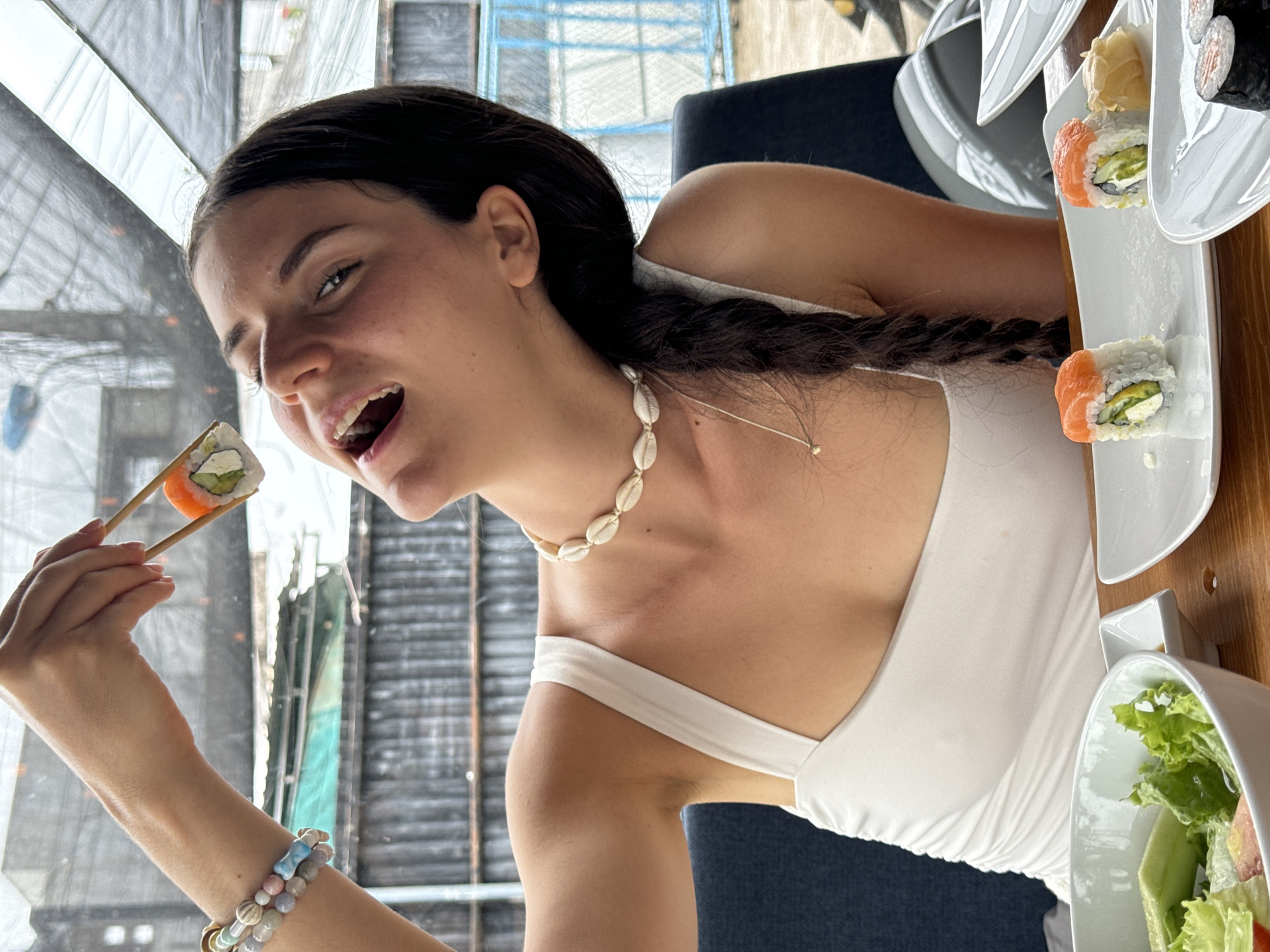 The image captures a woman at a table eating sushi. She is fair-skinned with dark hair styled in a braid, wearing a white tank top and a shell necklace. She is holding a piece of sushi with chopsticks near her mouth. The table holds plates of sushi and a bowl of salad. The location is Phu Thuy Ward, Vietnam. Through a window behind her, vertical structural elements can be seen.