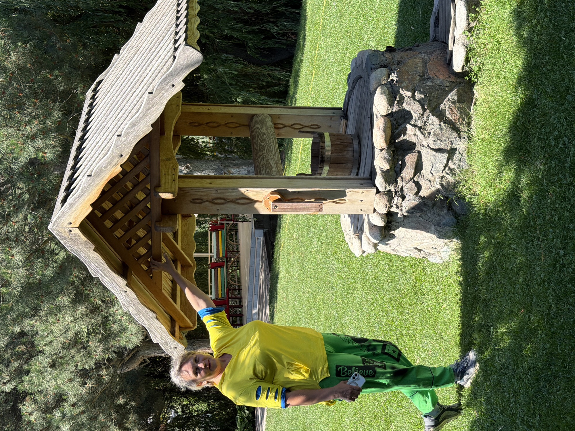 A woman with graying blonde hair, wearing a bright yellow t-shirt with blue accents on the sleeves and green pants with writing, stands to the left of a rustic wooden wishing well. She is holding a white mobile phone in her left hand and her right arm is raised, with her hand resting on the wooden structure of the well. The well has a pitched roof, wooden supports, a crank mechanism, and a wooden bucket inside. The well is constructed with a stone base and sits on a well-maintained green lawn. In the background, behind the well, are trees, and further back, colorful benches and outdoor seating are visible under a covered area. The scene is illuminated by sunlight, casting shadows on the grass.