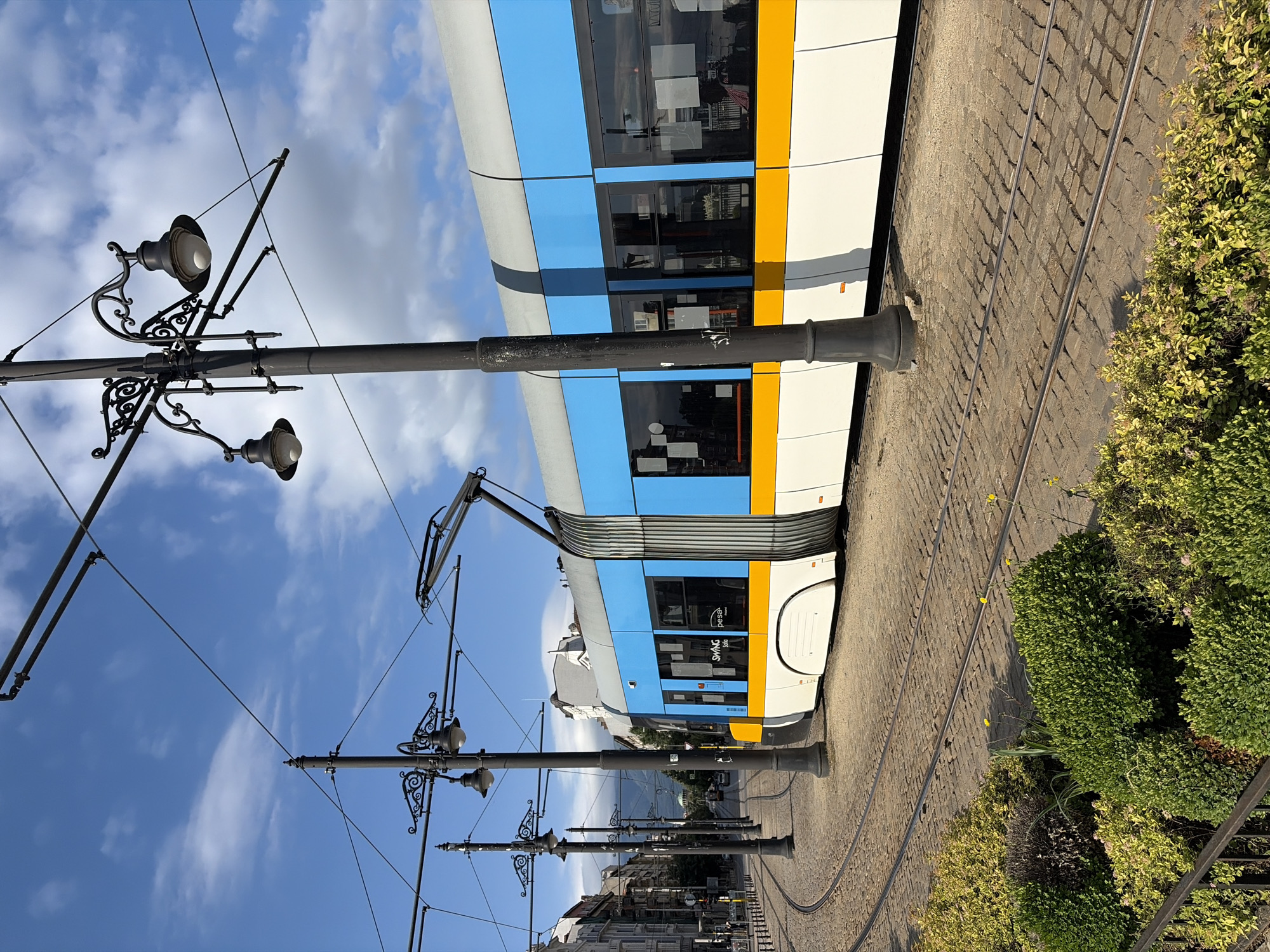 The image depicts a bright, sunny day in Sofia, Bulgaria, with a modern tram on a cobblestone street. The tram is predominantly blue and white with a yellow stripe. It's positioned on tram tracks that curve into the distance. Several ornate, dark lampposts line the street, providing a contrast to the modern tram. The sky is a vibrant blue with scattered white clouds. In the foreground, there are lush green bushes and a black metal fence. The background shows buildings typical of a European city, some with visible architecture details and windows. The overall scene is a blend of historical elements, like the cobblestones and lampposts, with contemporary urban transit.