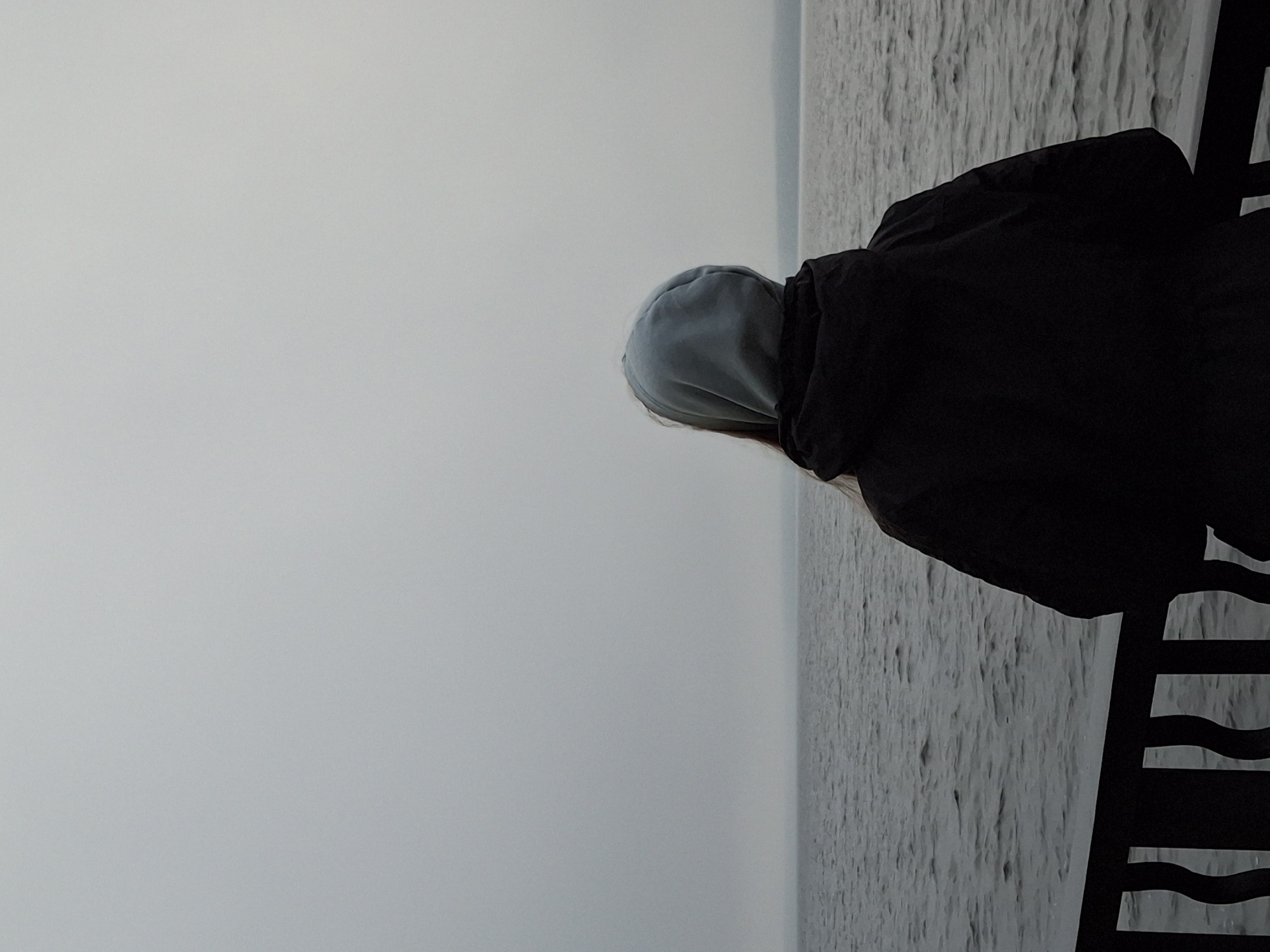A person stands facing away from the camera, looking out at a body of water under a cloudy sky. The person is wearing a light blue hooded sweatshirt over a dark jacket. They are positioned on a walkway or balcony with a dark railing in the foreground. The water appears calm with small ripples. The horizon is indistinct due to the fog or overcast conditions, with a faint line of land visible in the distance. This scene likely depicts a location along the Kama River, which flows through the city of Chaikovsky, Russia, possibly from a public embankment or viewing area.