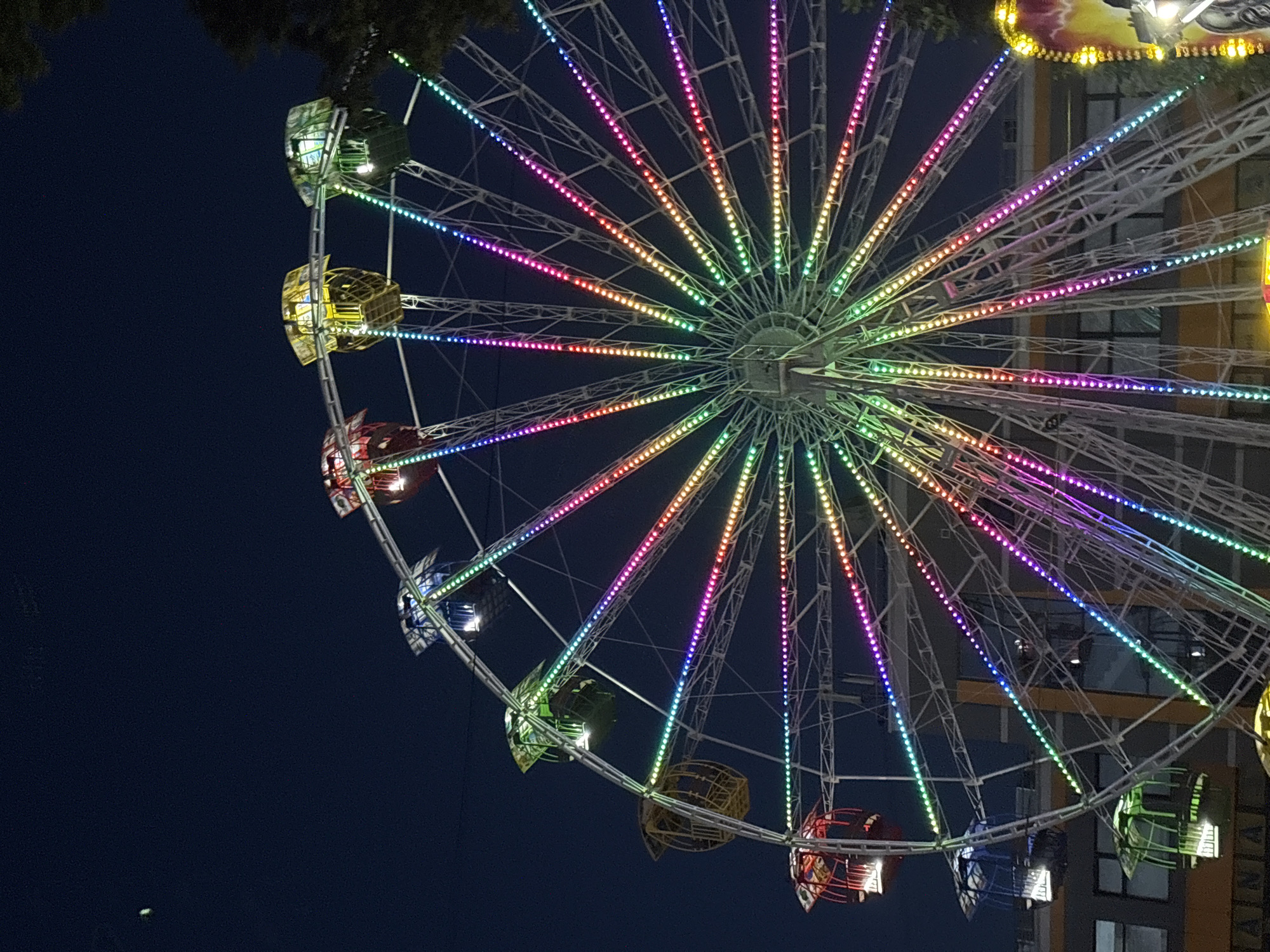 A Ferris wheel illuminated with colorful lights stands against a dark night sky. The Ferris wheel has multiple gondolas, each with a different colored roof. The lights on the spokes of the wheel transition through a spectrum of colors, including green, yellow, pink, and blue. In the background, the upper floors of a building are visible, with some windows illuminated. A portion of a tree is visible on the right side of the frame, and a smaller object, possibly the moon or a planet, is visible in the upper left quadrant of the sky. The scene appears to be at an amusement park or fairground in Bila Tserkva, Ukraine.