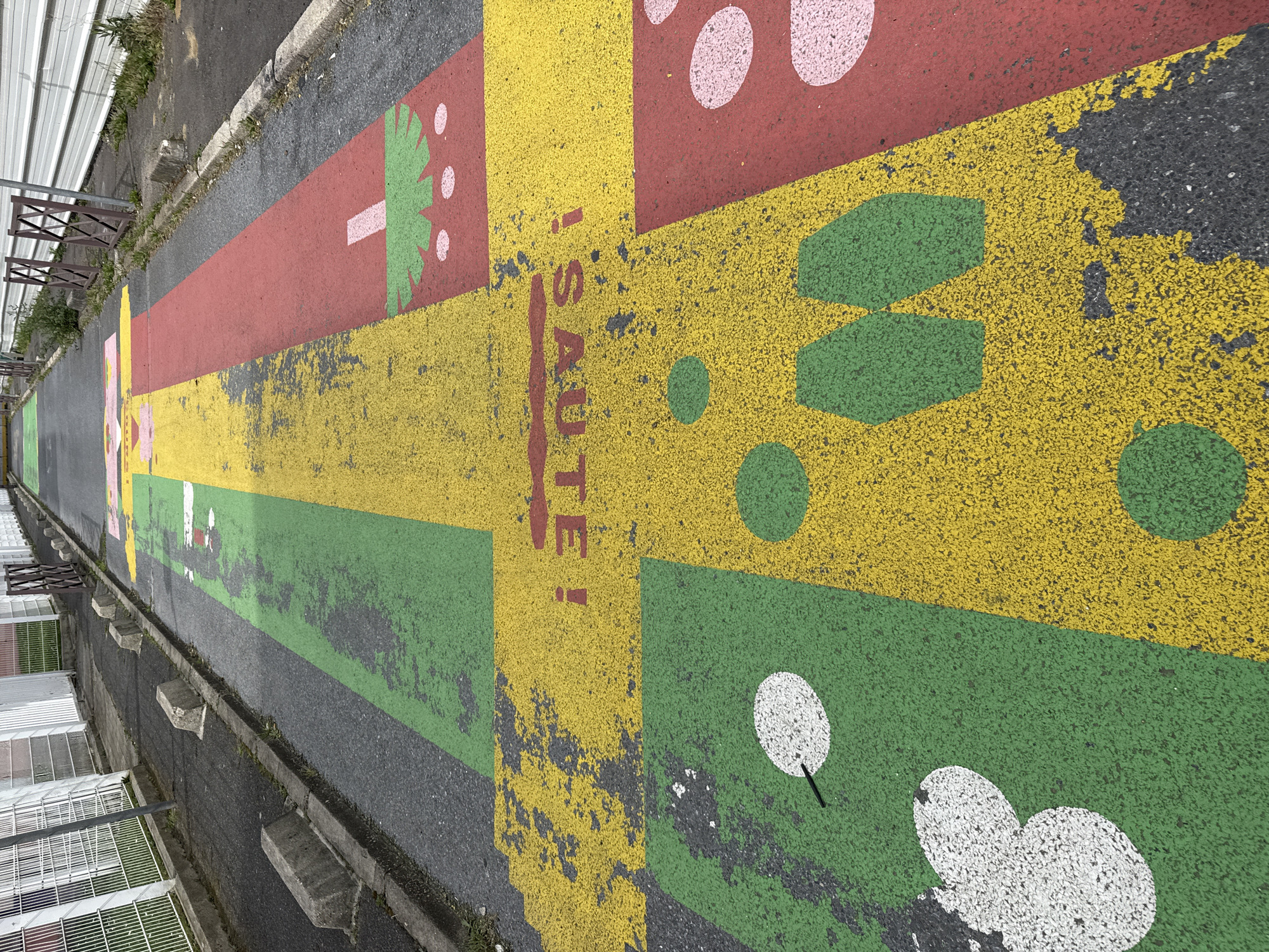 This image shows a section of an outdoor hopscotch game painted on asphalt. The game's design consists of various colorful rectangular sections in red, yellow, and green, each containing different shapes and patterns, including circles, handprints, and stylized depictions of plants. The word "SAUTE!" (meaning "jump!" in French) is painted vertically in red.
The asphalt surface shows signs of wear and tear, with some areas where the paint has faded or chipped away. The game is situated alongside a curb, and on the left of the frame, a building with metal grating is visible. This hopscotch game is located in Sarcelles, France, as identified by the context provided.