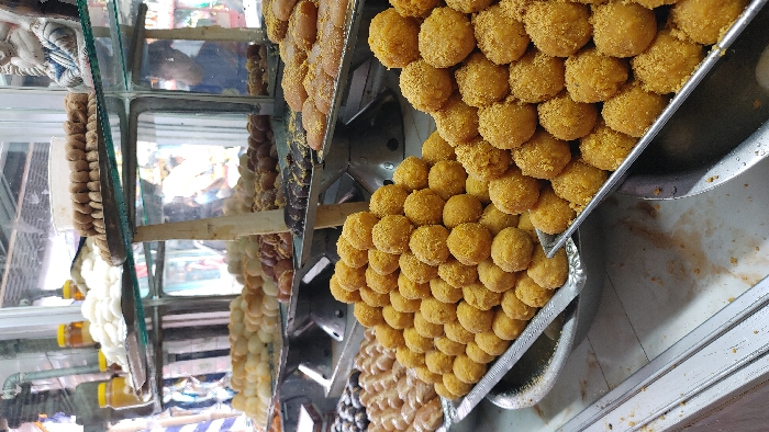 This image depicts an indoor commercial setting in Bandar, Bangladesh, specifically a sweet shop. The foreground is dominated by two large, mounded piles of yellow, spherical sweets, likely *ladoos*, arranged on rectangular silver-colored metal trays. These sweets feature a granular, lighter-colored coating. Behind these, a multi-tiered glass display case showcases various other confectionery items. On the lower shelves of the display, numerous types of sweets are visible: white, round sweets; darker, brown, and light-brown spherical or oval sweets, some resting on dark metal scales; and elongated, bar-shaped items. The top shelf holds several yellow-capped jars, presumably containing honey or syrup, alongside a small, light-colored decorative figurine or deity statue. A reflection of an individual wearing a blue shirt is discernible within the glass of the display case, indicating the presence of people in the vicinity. The counter surface in the immediate foreground is a light-colored, possibly tiled, material.