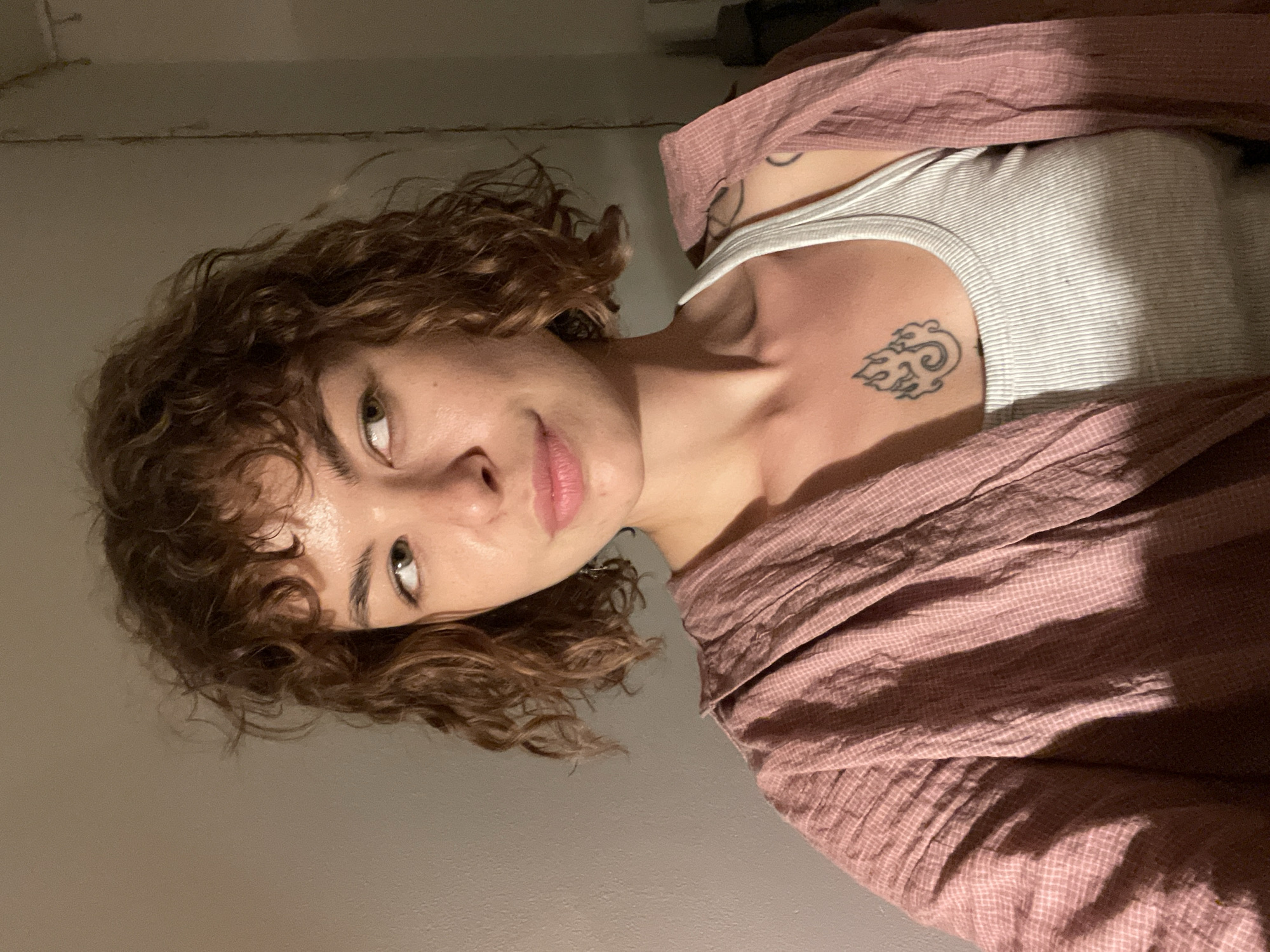 A woman with curly brown hair is looking at the camera. She is wearing a white tank top and a pink, checkered shirt draped over her shoulders. She has tattoos on her chest and shoulder. The background is a plain wall. The image does not provide sufficient information to verify a location in Boreti, Montenegro.