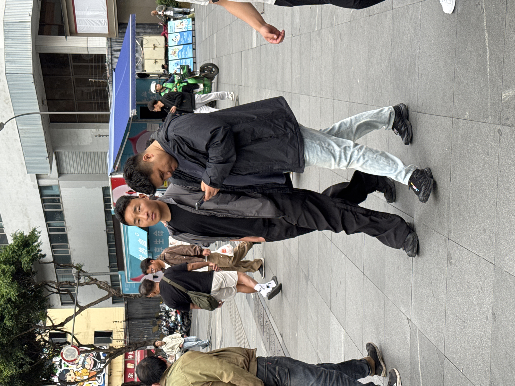 On a paved walkway in Jinjiang District, China, a group of pedestrians are present. Two men stand close together. One wears a black jacket and light blue jeans, the other a grey jacket and black pants. A man in shorts and a black shirt walks nearby, and another man in a tan jacket lies on the ground. Trees and buildings are visible in the background. There are motorcycles parked on the side of the road. The scene seems to depict an urban street scene with diverse people.