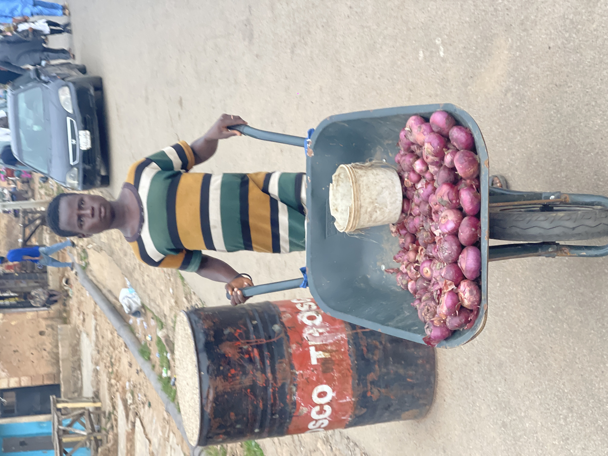 A man in Jos, Nigeria is pulling a wheelbarrow filled with red onions and a small, white container. He is wearing a striped shirt. Next to the wheelbarrow is an old black and red barrel with white lettering. A car is parked on the street in the background. The surrounding area appears to be a street with buildings and other urban elements.