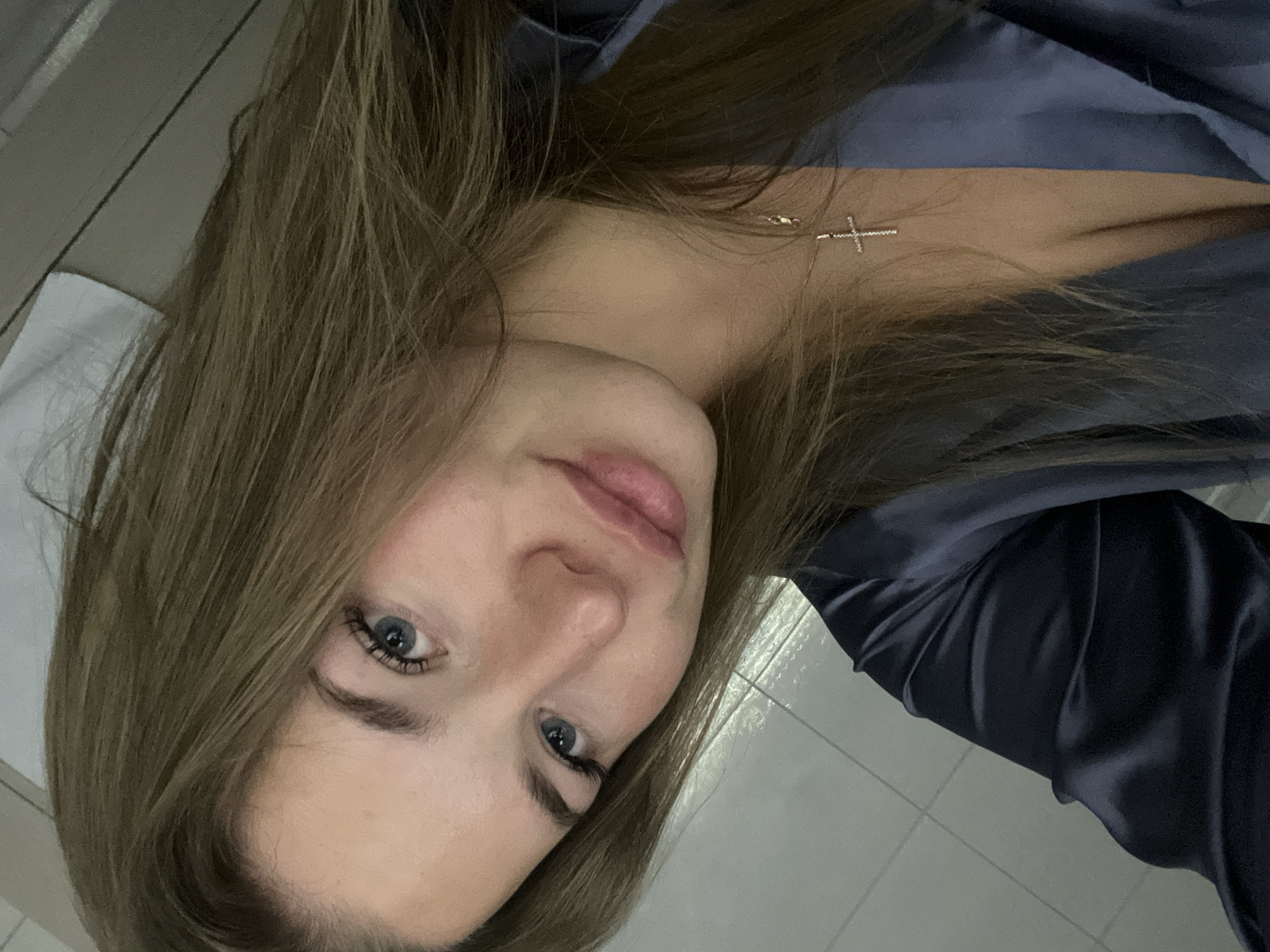 A young woman with long, light brown hair and blue eyes is posing for a selfie. She is wearing a dark-colored, possibly satin, top or robe, and a delicate silver cross necklace is visible on her chest. The background appears to be a bathroom, indicated by the white tiled walls and ceiling. The photo's orientation is tilted to the side. The location in the city of Budva, Montenegro, cannot be determined from the visual information.