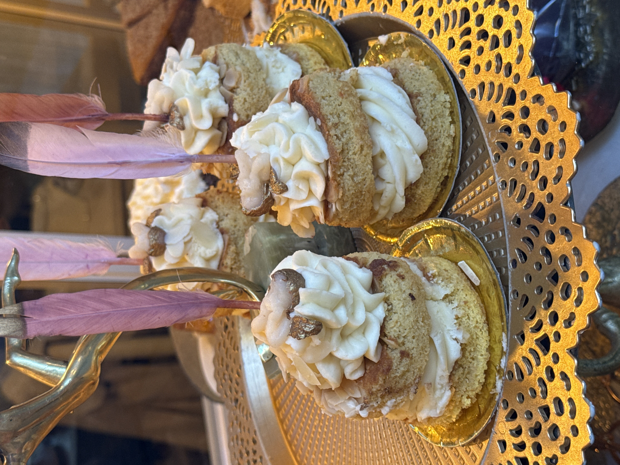 The image shows a display of ornate desserts, potentially in a shop window. Three sponge cake rolls filled with white frosting are arranged on individual gold-colored cups, which are placed on a decorative gold-colored tray with a perforated pattern around the edge. Pink feathers are inserted into the sides of the desserts, possibly for decorative purposes. The desserts appear to be topped with small pieces of gold leaf. Based on the style of the desserts and decor, this image could be from a shop in Barcelona, Spain, known for its pastry shops and ornate displays. No people are visible in the frame. The focus is on the presentation and detail of the desserts and their arrangement.