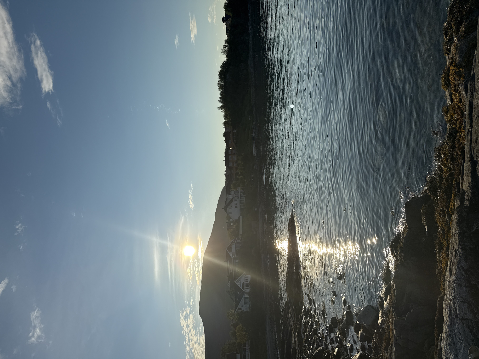 The image depicts a serene coastal landscape with a bright sun positioned above a silhouetted mountain range in Harstad, Norway. The sun's rays reflect off the gently rippling water, creating a shimmering effect. Several houses are visible nestled on the hillside, surrounded by trees. The foreground shows a rocky shoreline covered with some vegetation. The sky is a clear blue with a few wispy clouds, suggesting a pleasant day. A small, bright circular object, possibly a buoy or a reflection, is visible in the water. The overall scene conveys tranquility and natural beauty.