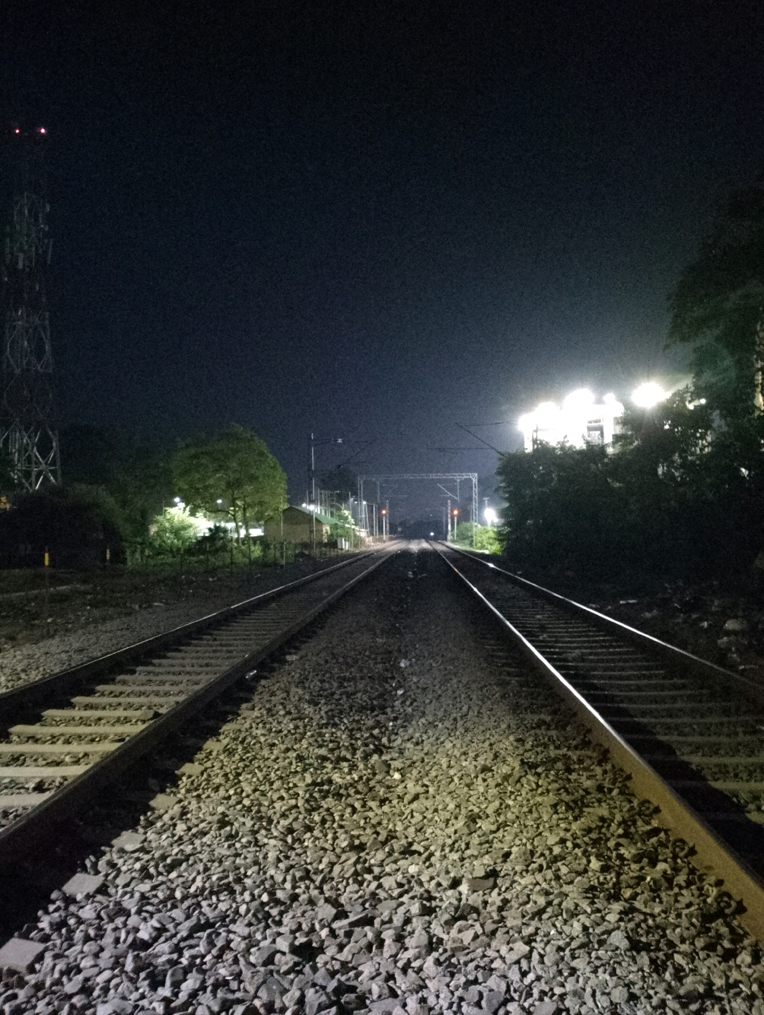 This is a night photograph of a railway track in Maligaon, India. The perspective is from ground level, looking down the parallel tracks which converge towards a distant point. The tracks are laid on a bed of gravel, illuminated by overhead lights.
To the left, a tall transmission tower with red lights at the top is visible in the distance. Further down the track, there are trees and a building structure with bright lights illuminating the area. On the right side, dense foliage and trees partially obscure the view, with more bright lights suggesting buildings or infrastructure. The sky is dark and overcast. No people are present in the image. The overall impression is one of a quiet, industrial night scene at a railway junction.