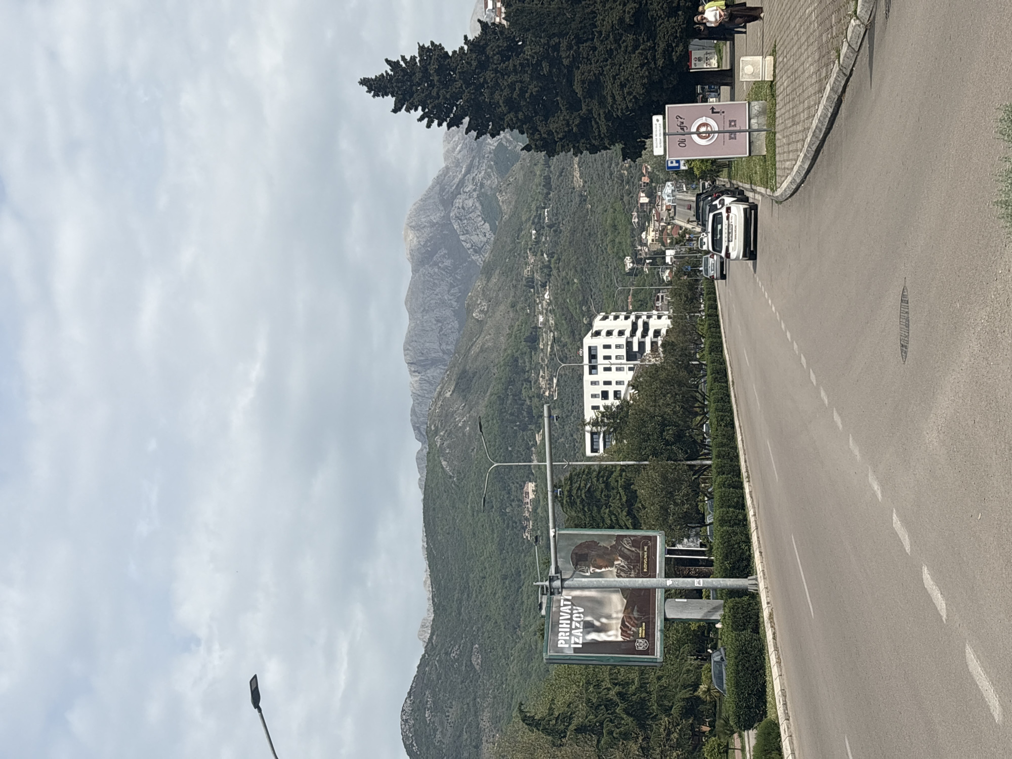 The image depicts a daytime scene in Bar, Montenegro, showcasing a road that runs parallel to a mountainous landscape under an overcast sky. The asphalt road features white dashed lines, and a paved sidewalk borders it on the left. Several cars, including a white SUV, are parked along the side of the road, and a person is visible walking on the sidewalk near the SUV.
In the foreground, two street-level signs are present. On the left side of the road, a billboard features an image of a soldier with text in a Slavic language, reading "PRIHVATI IZAZOV" (Accept the Challenge) and "BUDIVOJNE.ME," likely a military recruitment advertisement. Further down on the right, another sign displays a washing machine icon and partially visible text "Oš Lju?" and "084/?".
The mid-ground reveals a landscape of verdant, tree-covered hillsides, dotted with buildings, including a distinctive modern white multi-story building with dark windows. A tall, dark conifer tree stands prominently in the upper right quadrant of the image. The background is dominated by towering, rugged mountains with rocky outcrops, their distant peaks appearing muted under the cloudy sky. The overall scene suggests a blend of urban infrastructure with dramatic natural scenery, characteristic of a coastal town. The lighting indicates a bright, cloudy day.