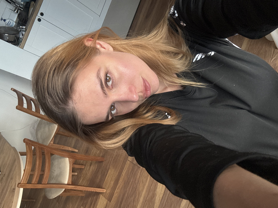 The image captures an indoor residential or kitchen area, location in the city of Unknown, Unknown. A female individual, appearing to be an adult, is centered in a close-up, selfie-style shot, looking directly at the camera. She has long, light brown hair with lighter highlights, parted slightly to the left and falling over her shoulders. Her complexion is fair, and she has light brown eyes with a neutral expression. She is wearing a black long-sleeved shirt, possibly athletic wear, featuring white triangular and hexagonal patterns on the shoulders and sleeves. Her right arm is extended, indicating she is holding the camera device.
In the background, to the subject's upper left, a light wooden dining table and two matching wooden chairs with light-colored upholstered seats are visible on a reddish-brown wood plank floor. To the subject's upper right, a portion of a kitchen area is visible, featuring white upper cabinets with black circular handles. On the counter beneath the cabinets, a stainless steel pot or pan, a white plastic dispenser, and part of a silver faucet are discernible. The walls in the background are light-colored. The scene is illuminated by ambient light.
