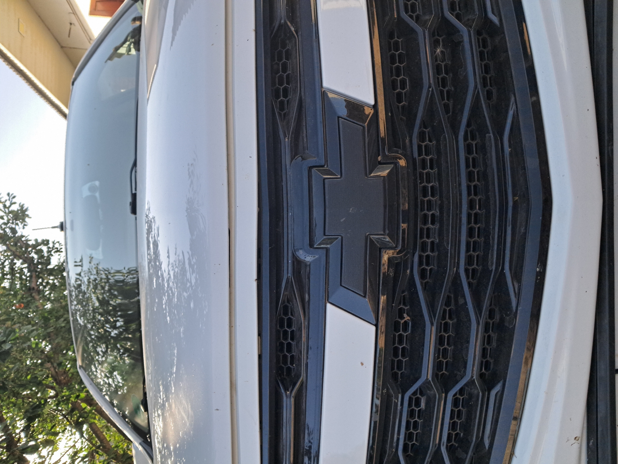 This is a close-up, low-angle, medium shot of the front grille of a white Chevrolet vehicle. The grille is black with a honeycomb pattern and features the prominent black Chevrolet bowtie emblem in the center. The hood of the car is visible above the grille, reflecting the sky and trees. The background shows a blurry view of a building with a light-colored roof and some green foliage, suggesting an outdoor setting in what appears to be a residential or street area. The overall impression is of a vehicle parked outdoors, possibly in a city or town.