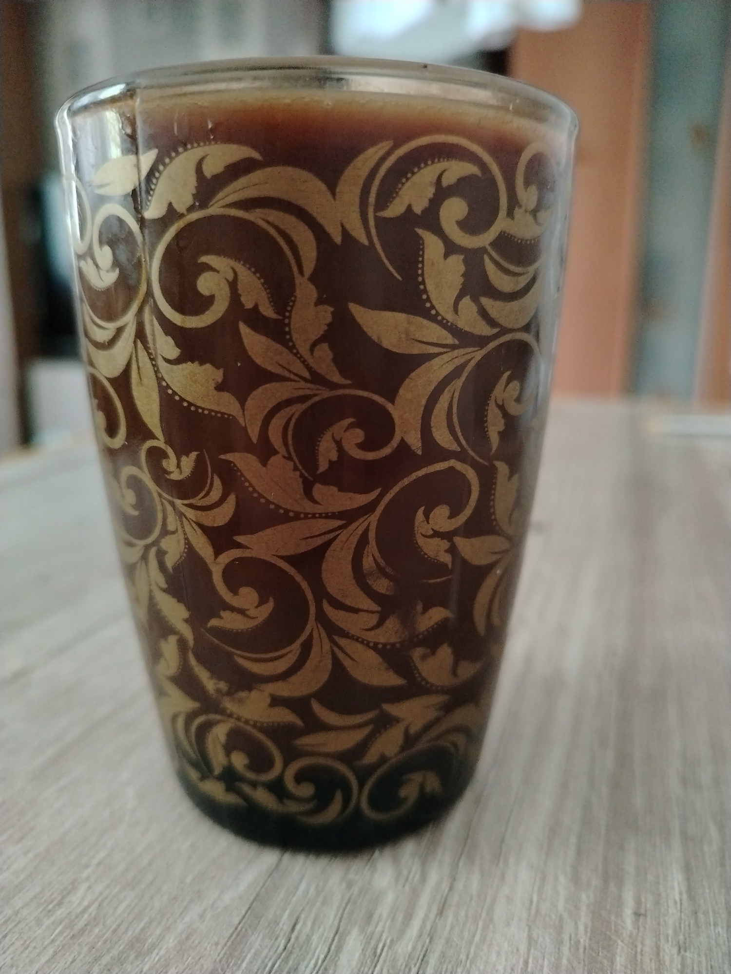 A close-up, eye-level shot features a dark brown glass tumbler with an ornate gold filigree pattern. The tumbler is filled with a dark liquid, possibly coffee or tea, and is positioned on a light-colored wooden table. The background is blurred, showing hints of furniture and a pale wall, suggesting an indoor setting. The image appears to be taken in a domestic environment, with no identifiable elements placing it specifically within Stavropol, Russia.